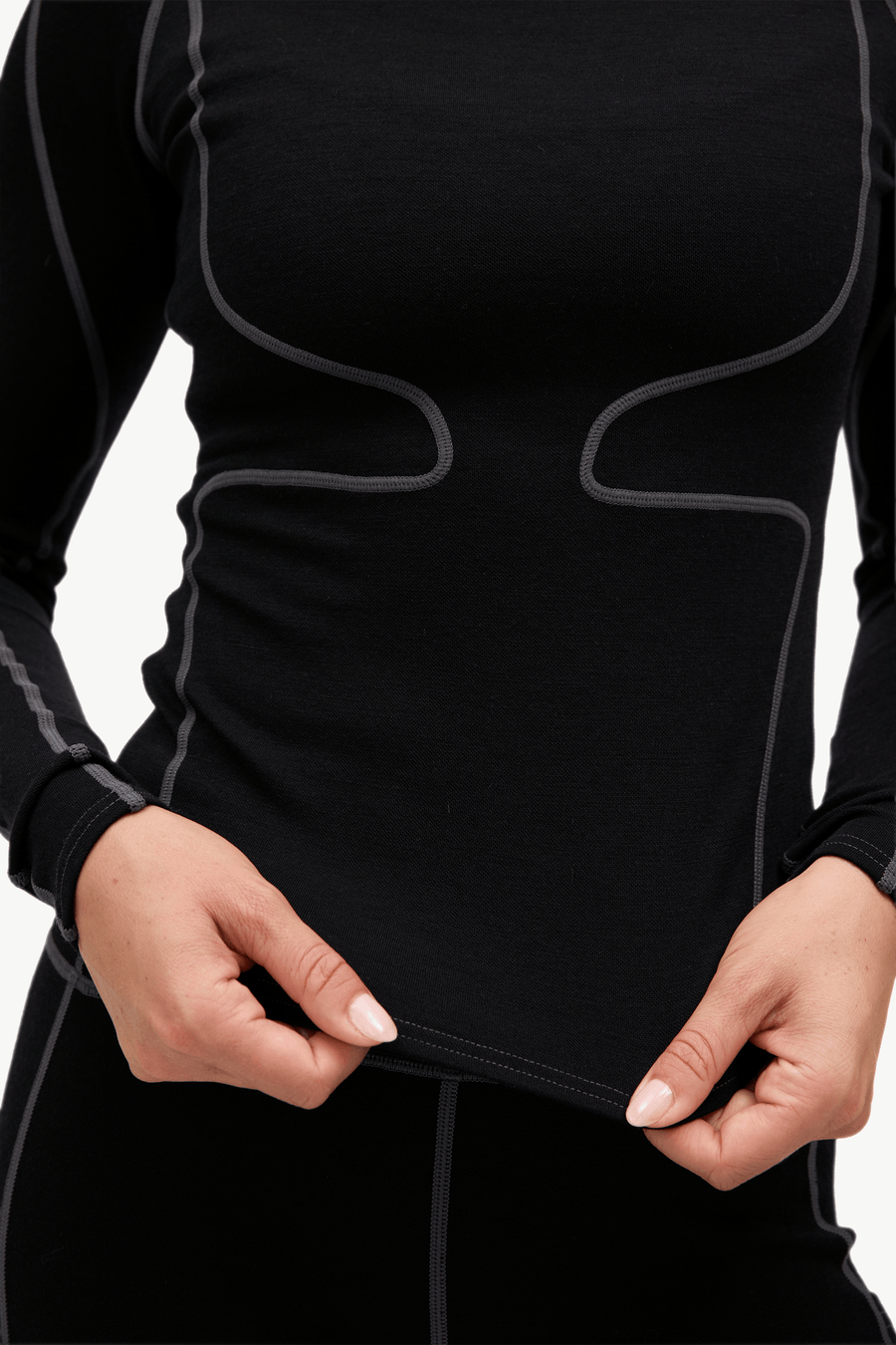 Close-up of a person wearing a black long-sleeve athletic compression top with gray contrast stitching, holding the hem of the shirt to show elasticity.