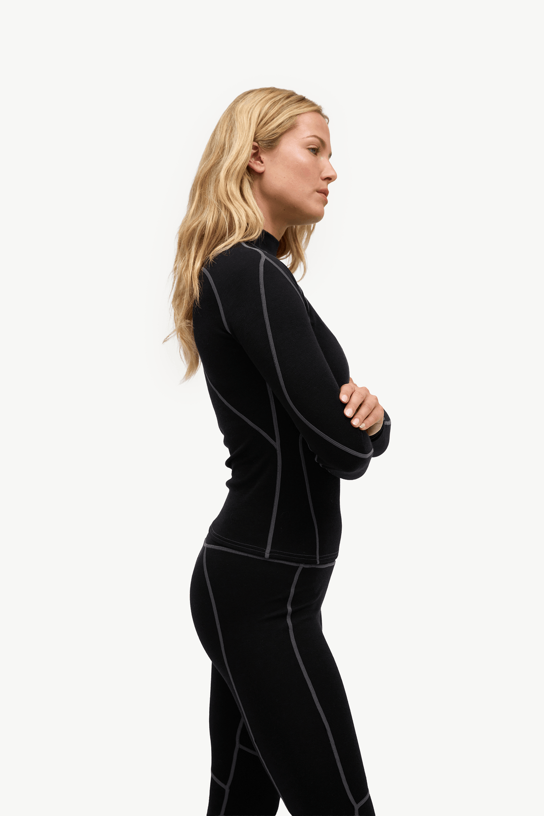 A blonde woman in profile wearing a black long-sleeve athletic top and matching black leggings with light gray seam detailing, posed against a solid off-white background.
