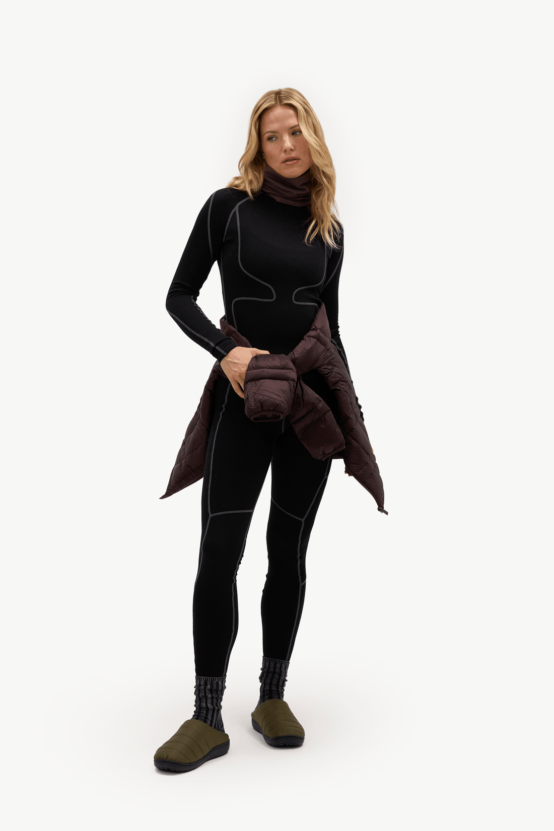 A woman modeling a black full-body wetsuit with contrast stitching, holding a dark brown puffer jacket, against a plain white studio background.