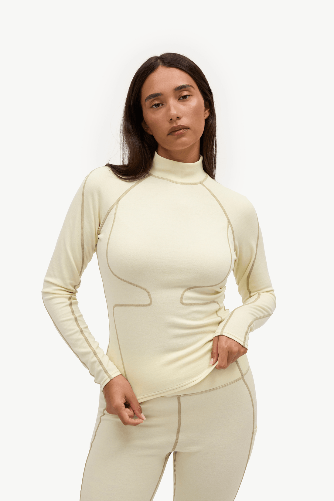 A woman modeling a fitted, long-sleeve mock-neck athletic top in a light cream color with contrasting light brown piping details, paired with matching light beige leggings, against a plain white studio background.