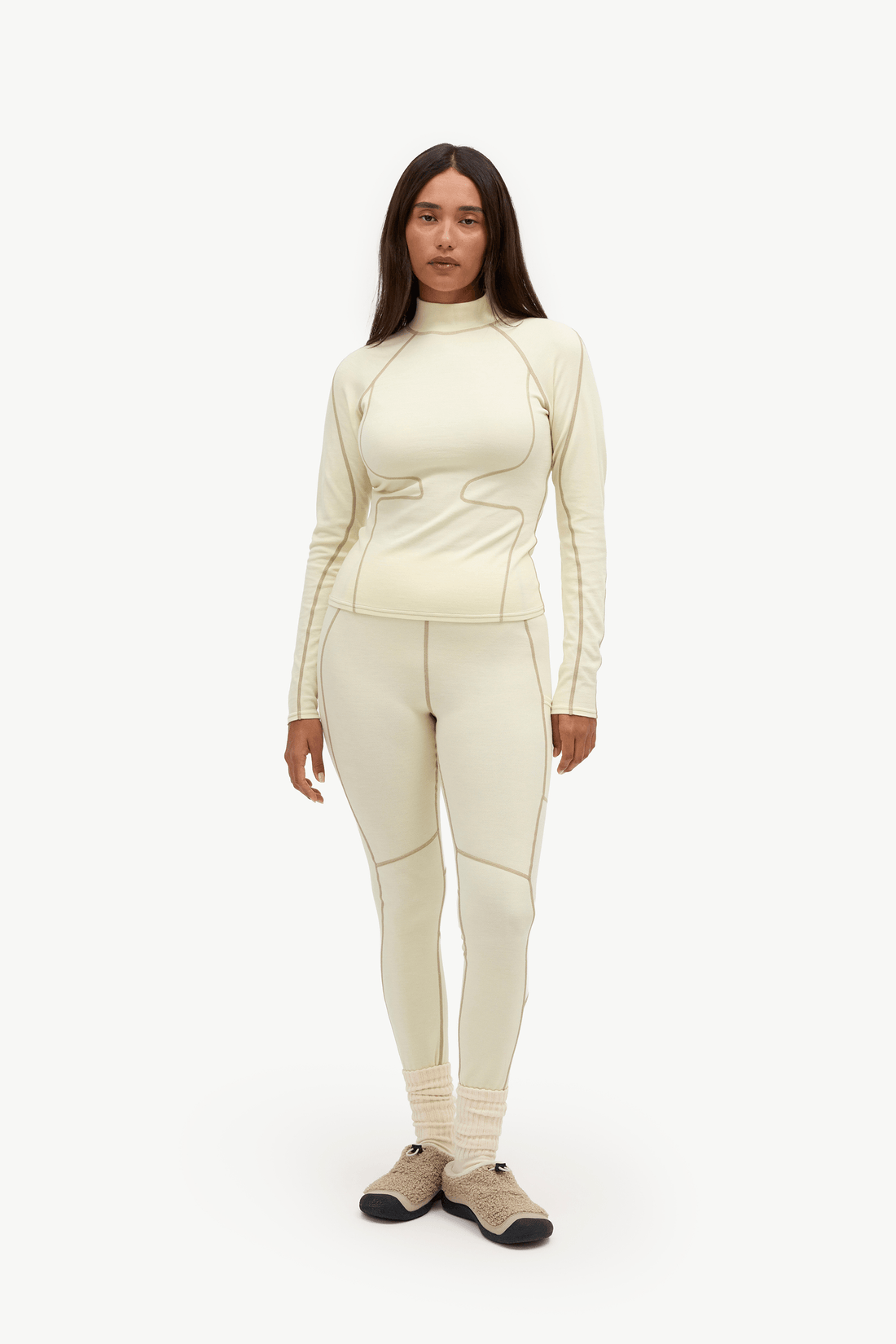 A woman modeling a matching cream-colored athletic set consisting of a long-sleeve mock-neck top and high-waisted leggings, paired with beige and black shoes, against a plain white studio background.