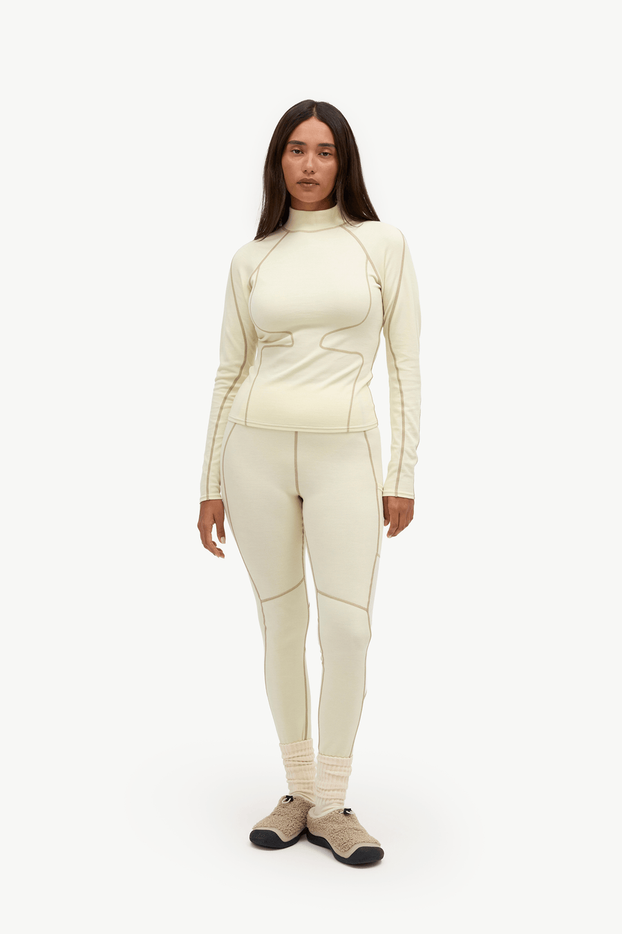 A woman modeling a matching cream-colored athletic set consisting of a long-sleeve mock-neck top and high-waisted leggings, paired with beige and black shoes, against a plain white studio background.