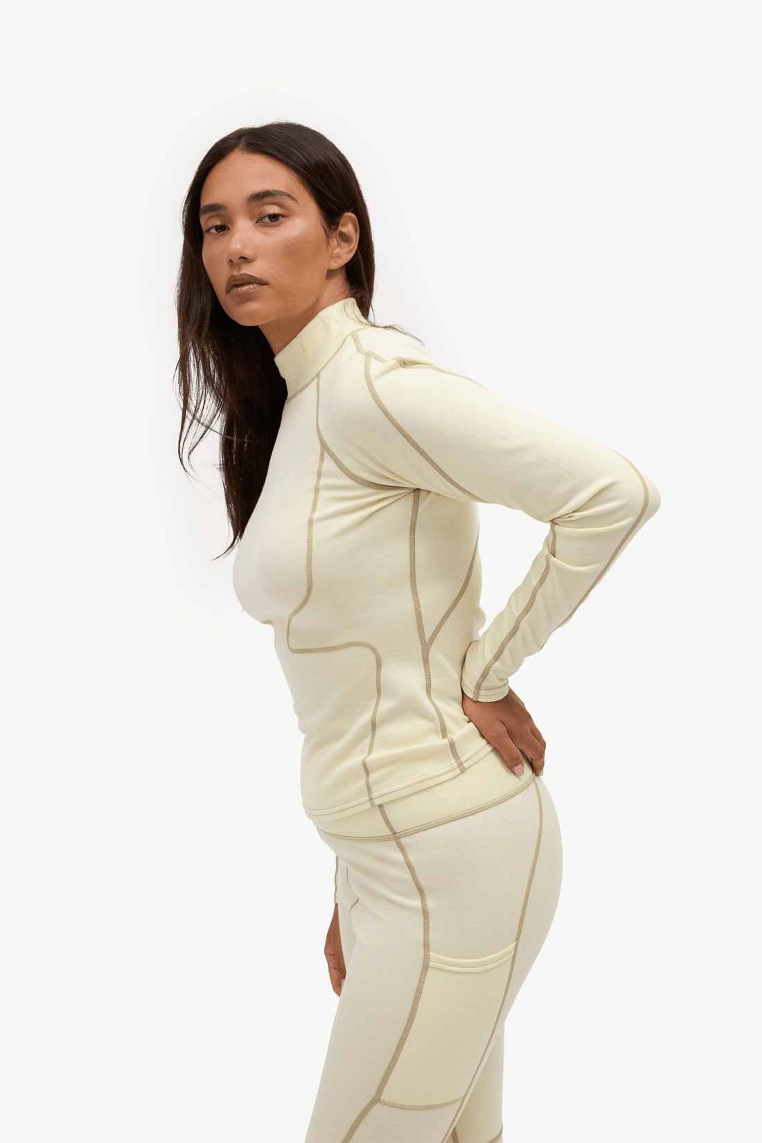 A woman modeling a matching cream-colored athletic set consisting of a long-sleeve mock-neck top and high-waisted leggings, featuring contrasting tan seam lines, against a plain white studio background.