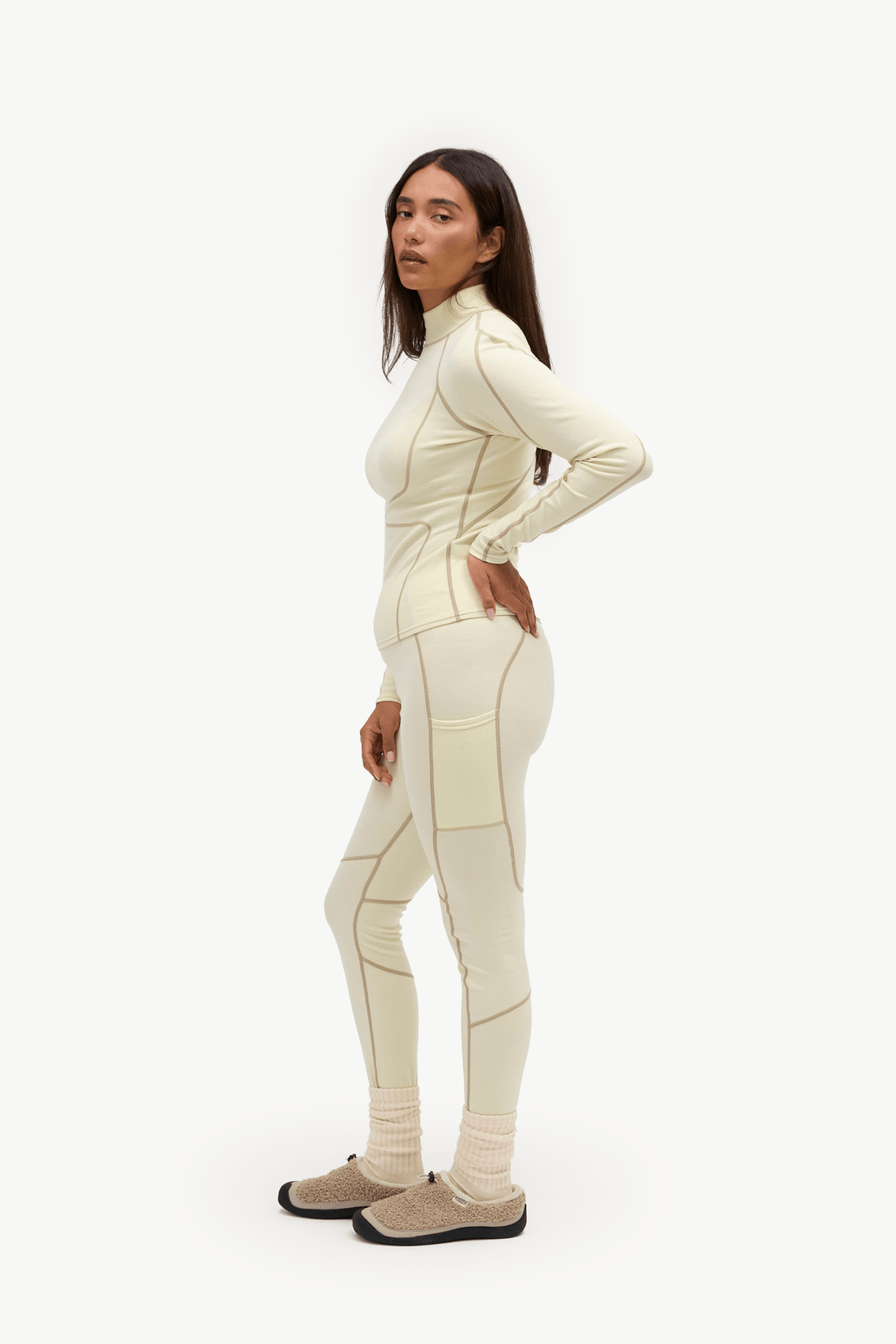 A woman modeling a matching cream-colored athletic tracksuit with beige contrast stitching, paired with beige knit socks and brown slip-on shoes, against a plain white studio background.