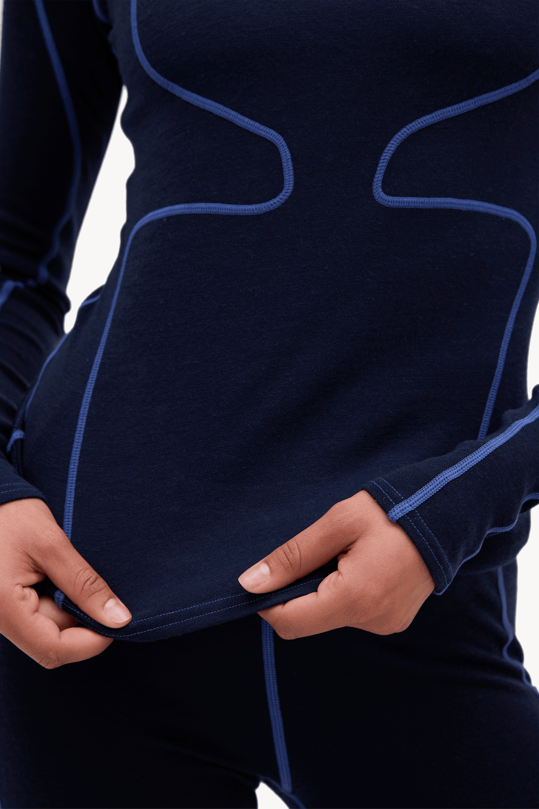 Close-up of a person wearing a dark navy long-sleeve athletic top with contrasting blue seam lines, holding the hem of the shirt to show flexibility. The background is solid white.