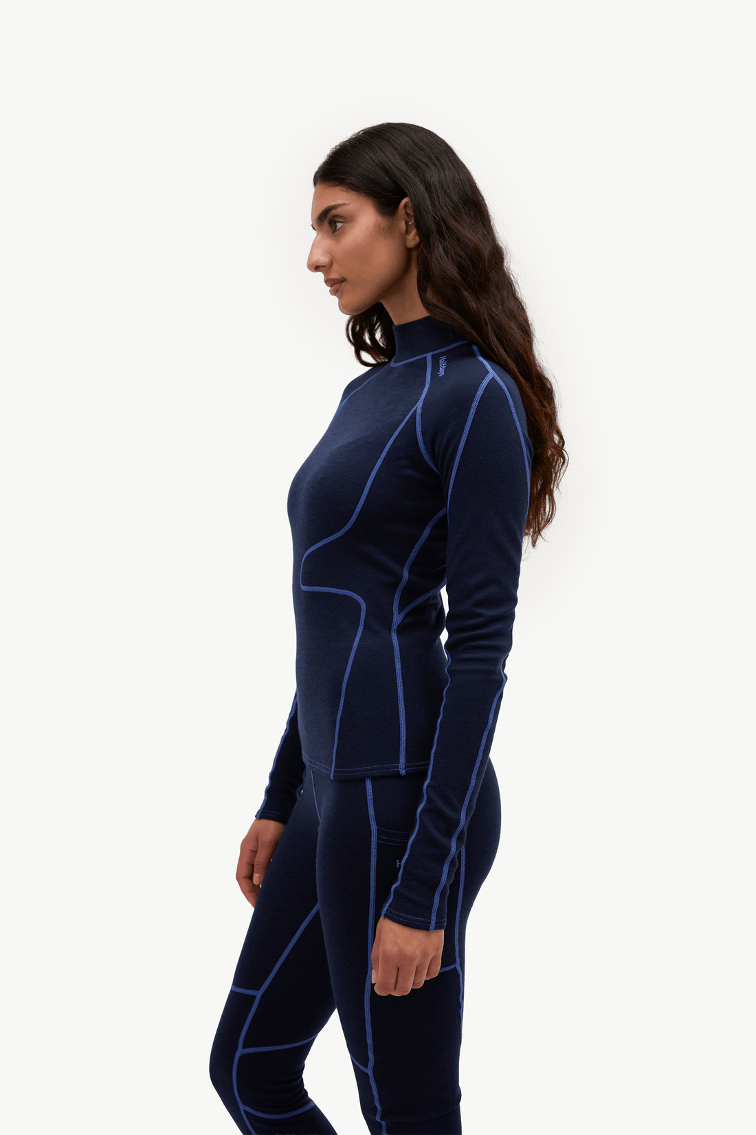 A woman in profile wearing a navy blue long-sleeve athletic top and matching leggings with bright blue contrast stitching, photographed against a clean white studio background.