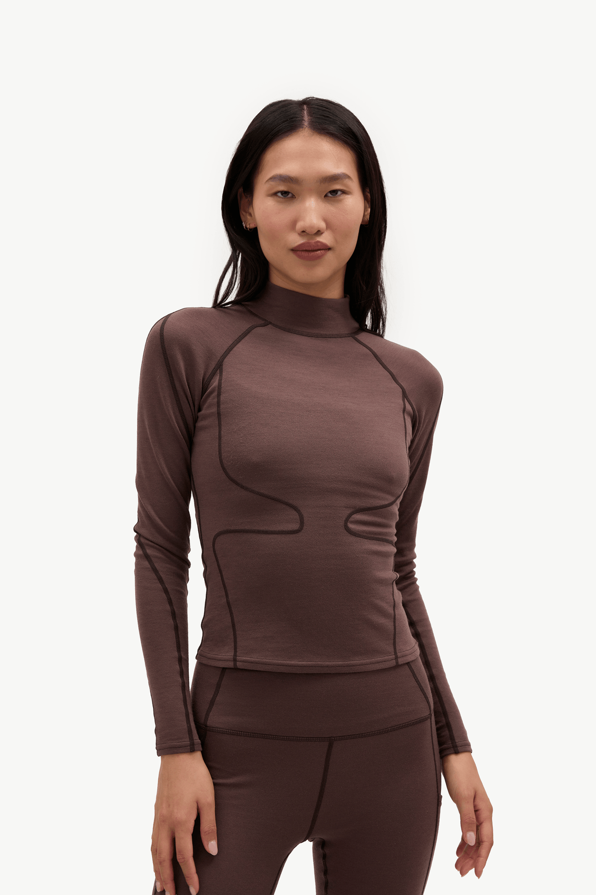 A woman modeling a fitted long-sleeve athletic top and matching leggings in a dark brown color, shown against a plain light gray studio background.