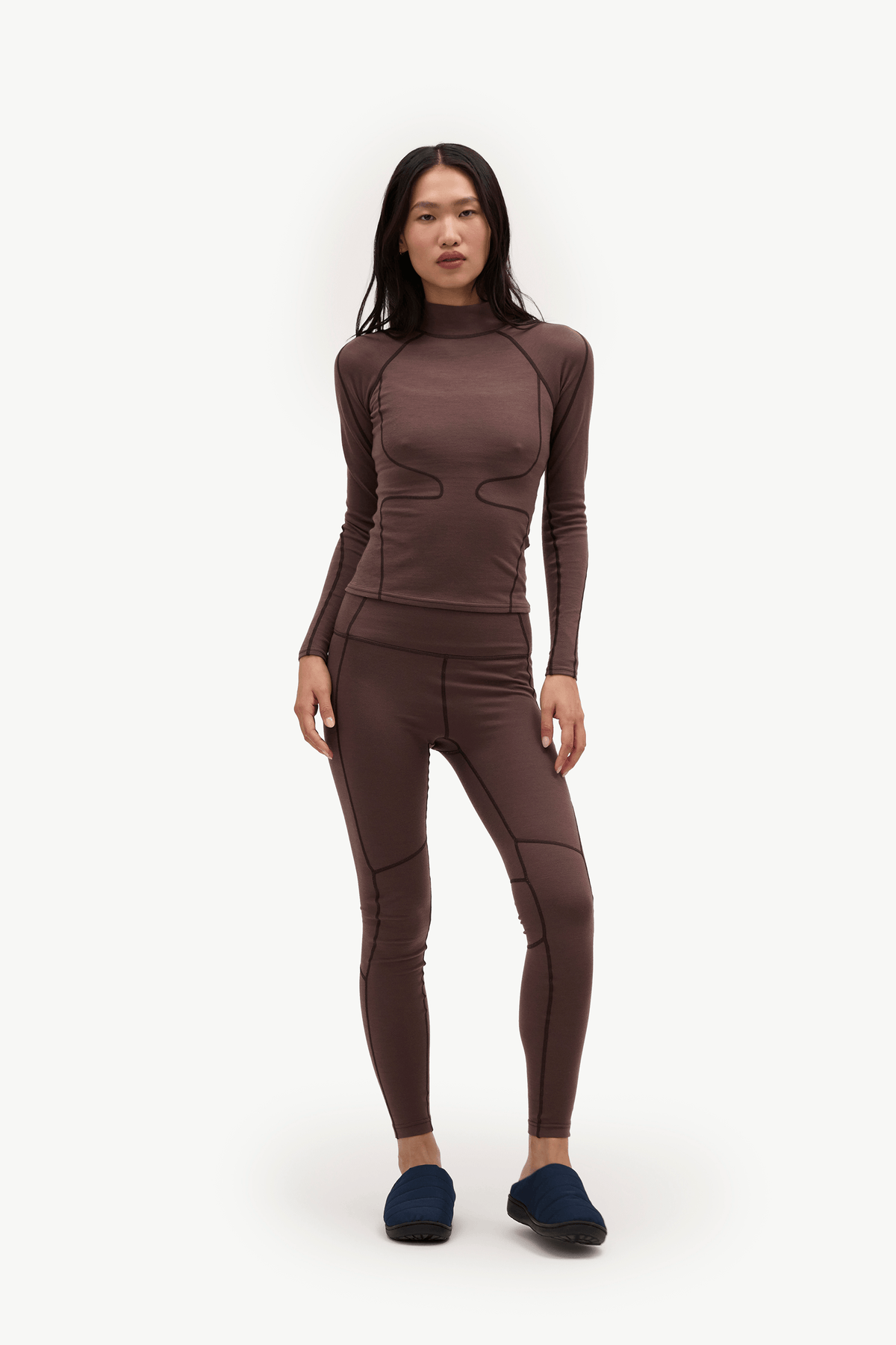 A woman modeling a matching dark brown long-sleeve mock neck top and high-waisted leggings set, paired with dark blue slippers, against a plain white studio background.