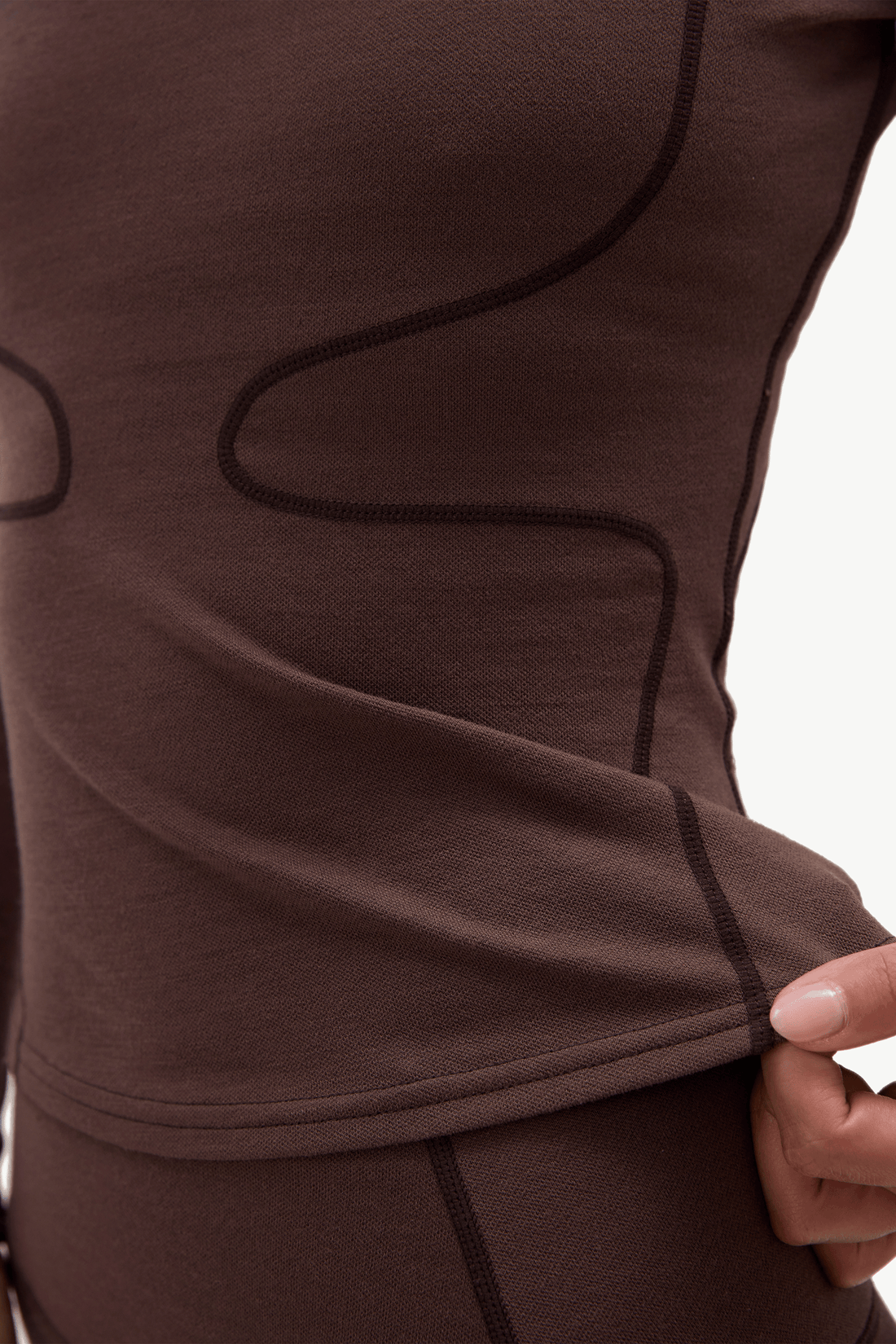 Close-up of a person wearing a brown athletic top, showing the side seam and a hand pulling the fabric to demonstrate stretch, against a plain white background.