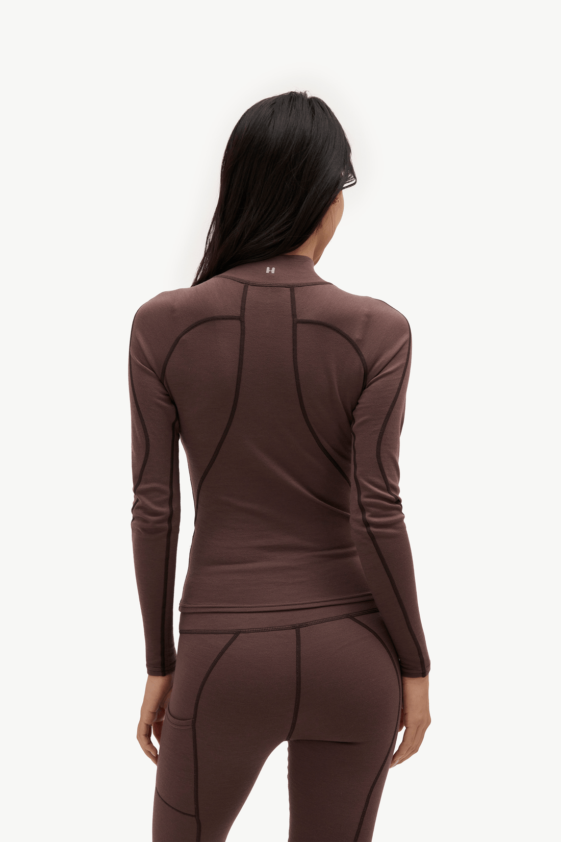 A woman viewed from the back wearing a fitted long-sleeve athletic top and matching leggings in a dark brown color, against a plain light background.
