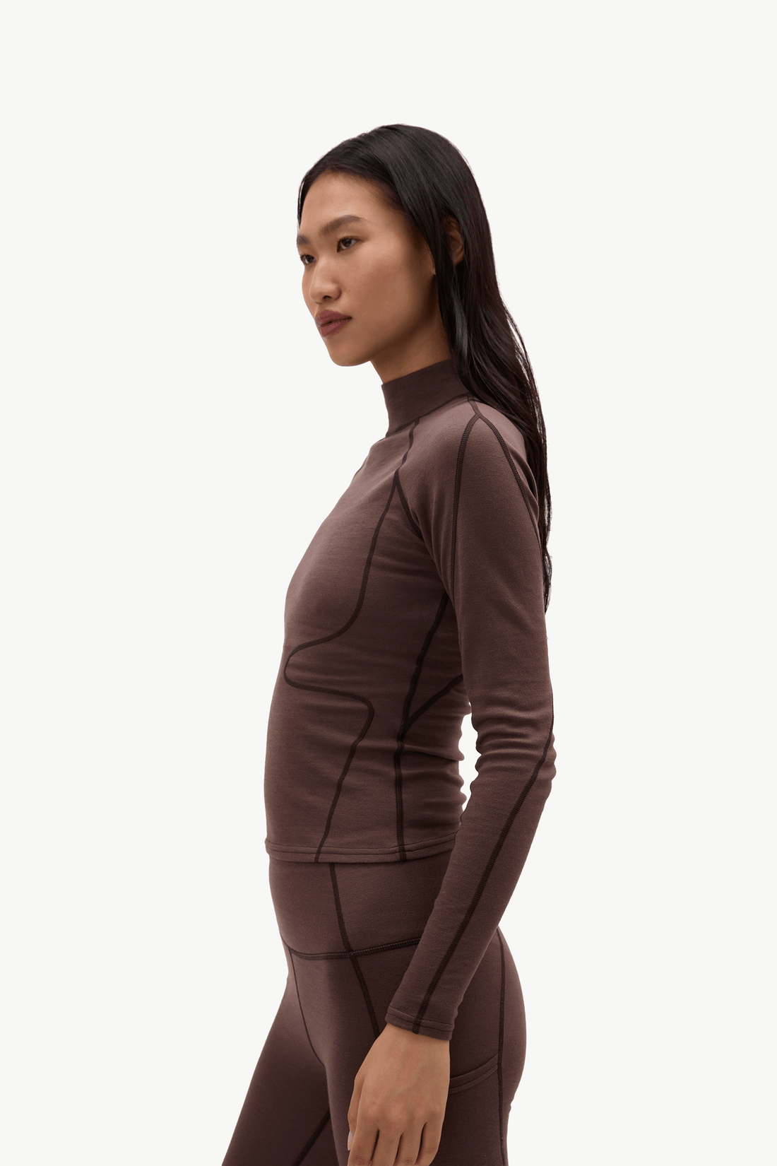 A woman in a side profile wearing a dark brown, form-fitting athletic long-sleeve top and matching leggings against a plain light background.