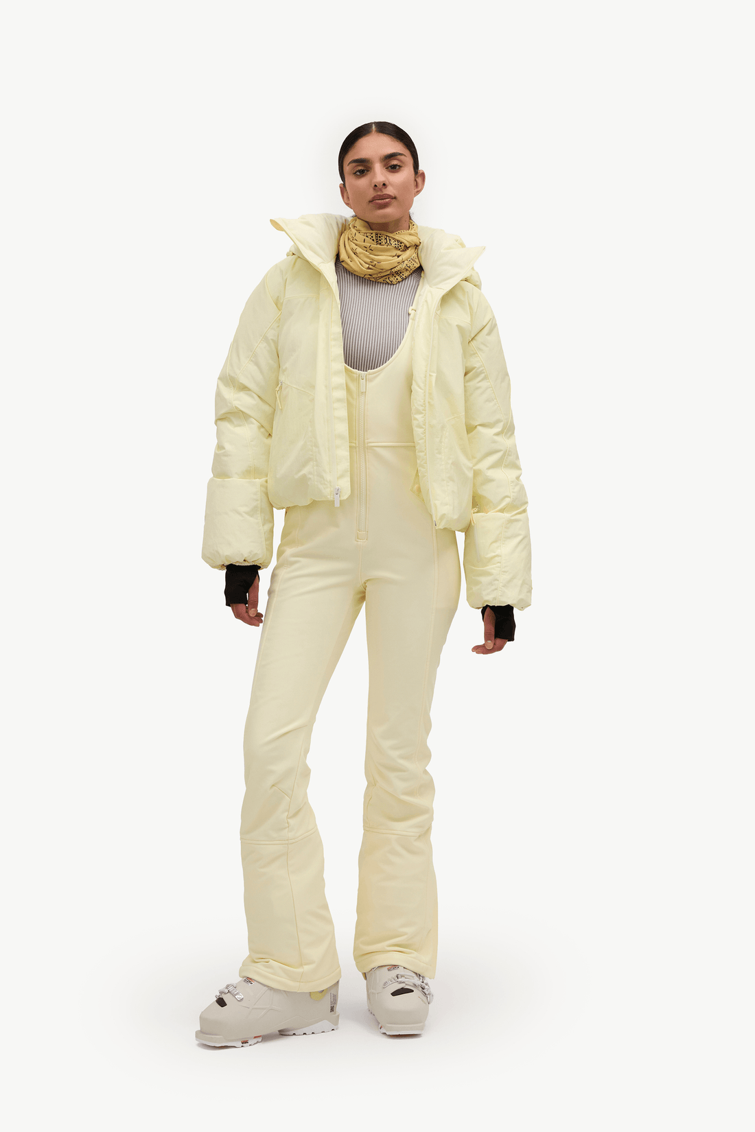 A model stands against a plain white studio background wearing a pale yellow puffer jacket, matching insulated pants, a gray top, a gold neck warmer, and light-colored sneakers.