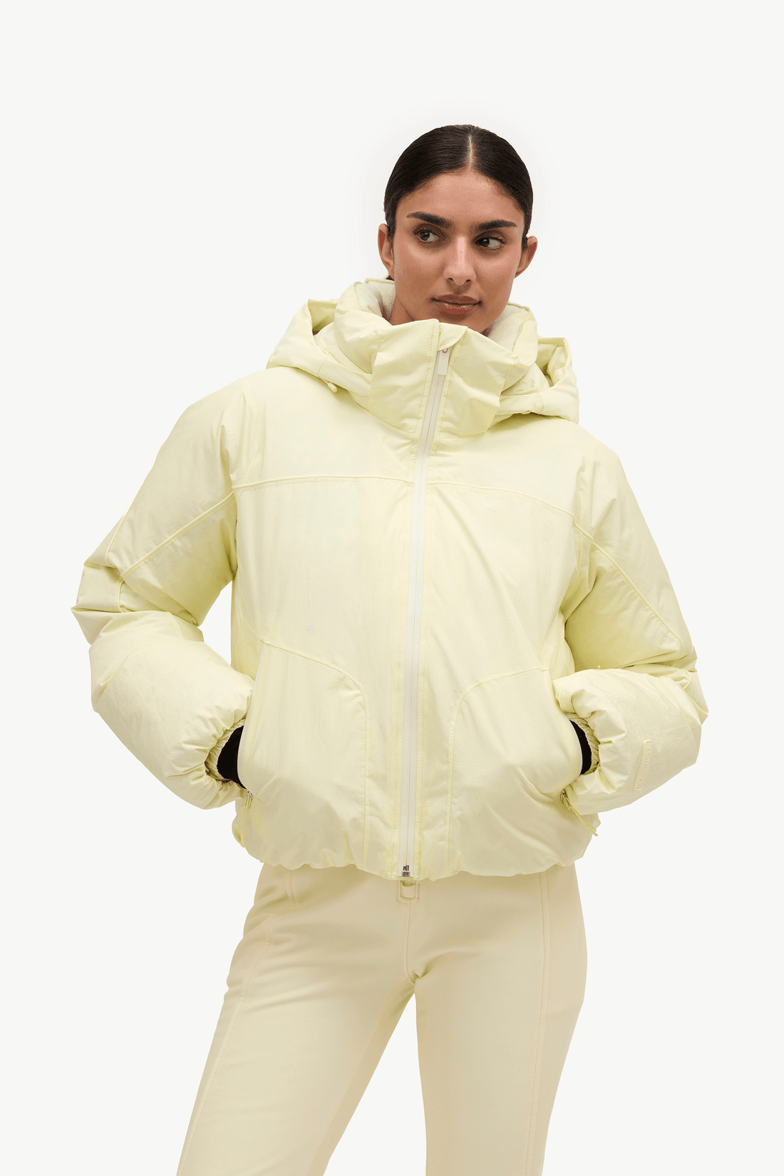 A model wearing a pale yellow puffer jacket with a high collar and front zipper, paired with matching light cream pants, against a plain white background.