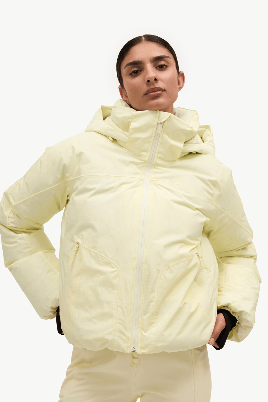 A model wearing a pale yellow puffer jacket with a high collar and front zipper, paired with matching light yellow pants, against a plain white background.