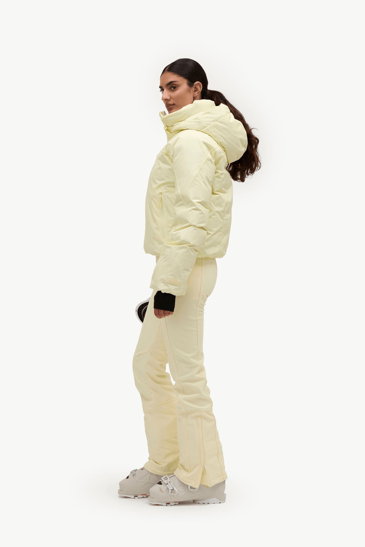 A woman modeling a pale cream puffer jacket and matching ski pants with light-colored snowboard boots, shown from a side profile against a plain white studio background.