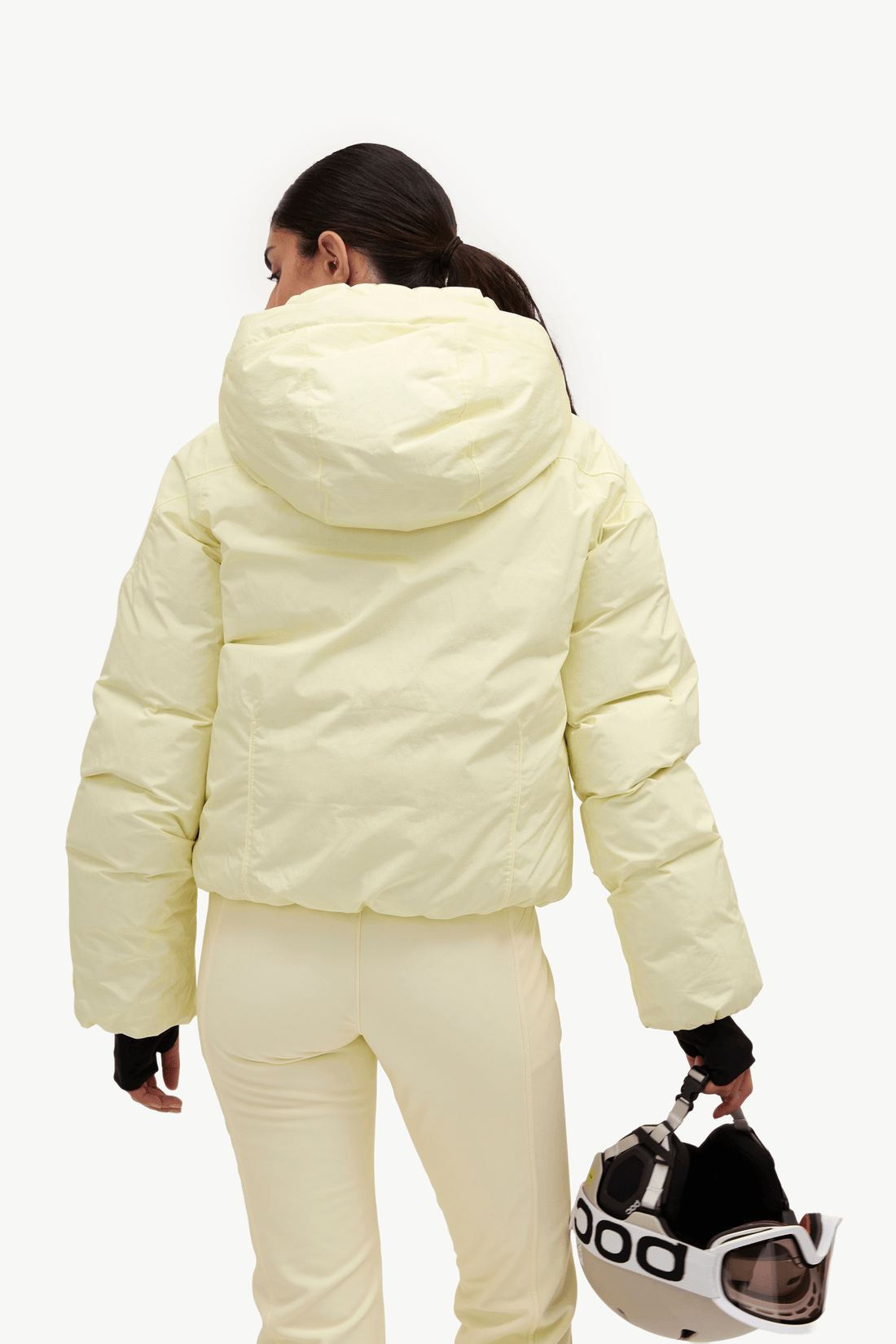 A woman shown from the back wearing a pale yellow puffer jacket and matching pants, holding a ski helmet against a plain light gray studio background.