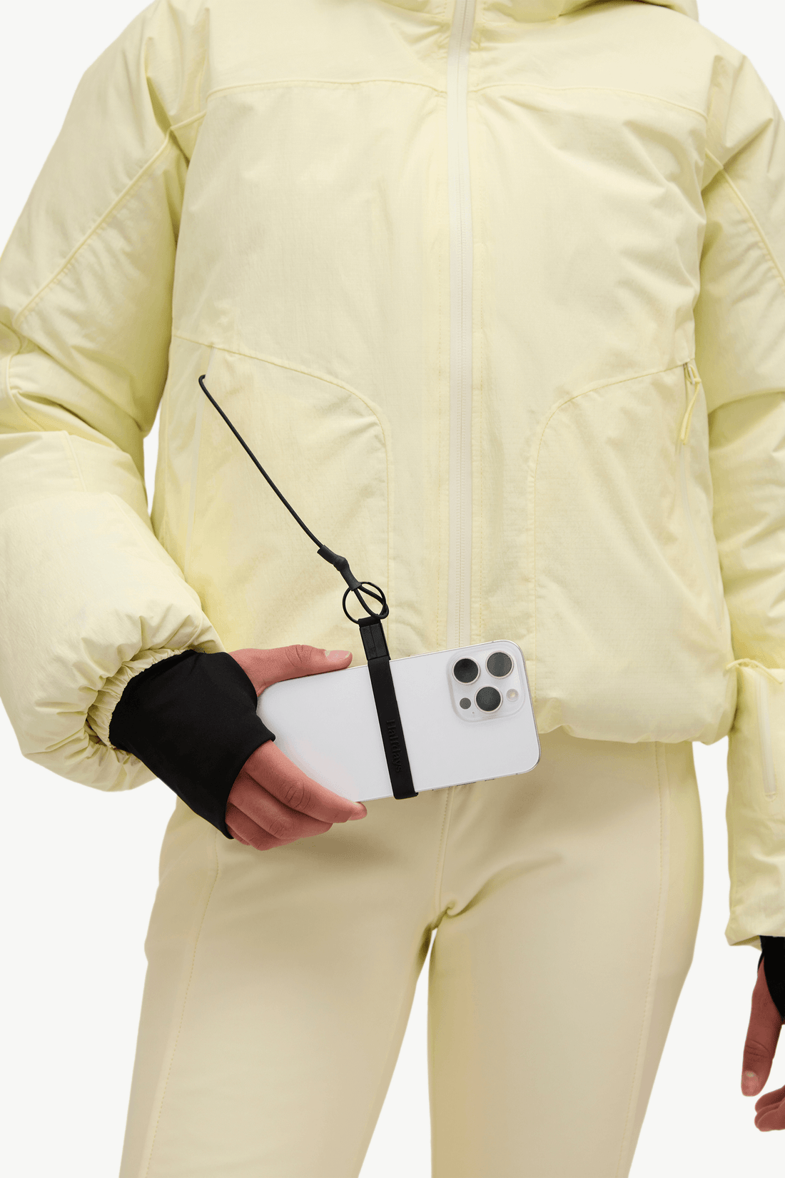 A close-up of a person wearing a pale yellow puffer jacket and matching pants, holding a white smartphone with a black wrist strap accessory attached near the sleeve.