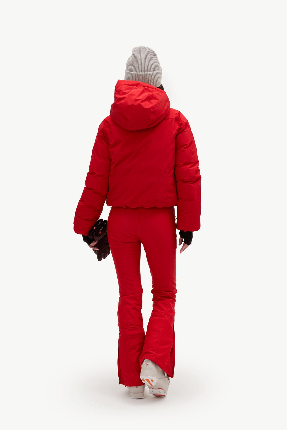 A model shown from behind wearing a bright red puffer jacket and matching red flared ski pants, a light gray knit beanie, and white sneakers, against a plain white studio background.