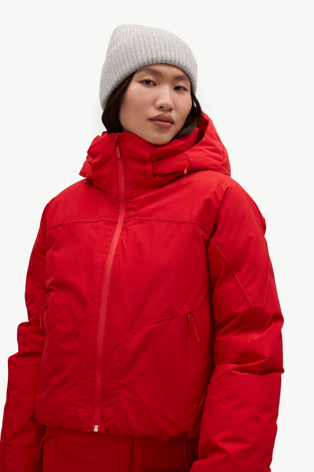 A model wearing a bright red hooded puffer jacket and a light gray knit beanie, photographed against a clean white studio background.