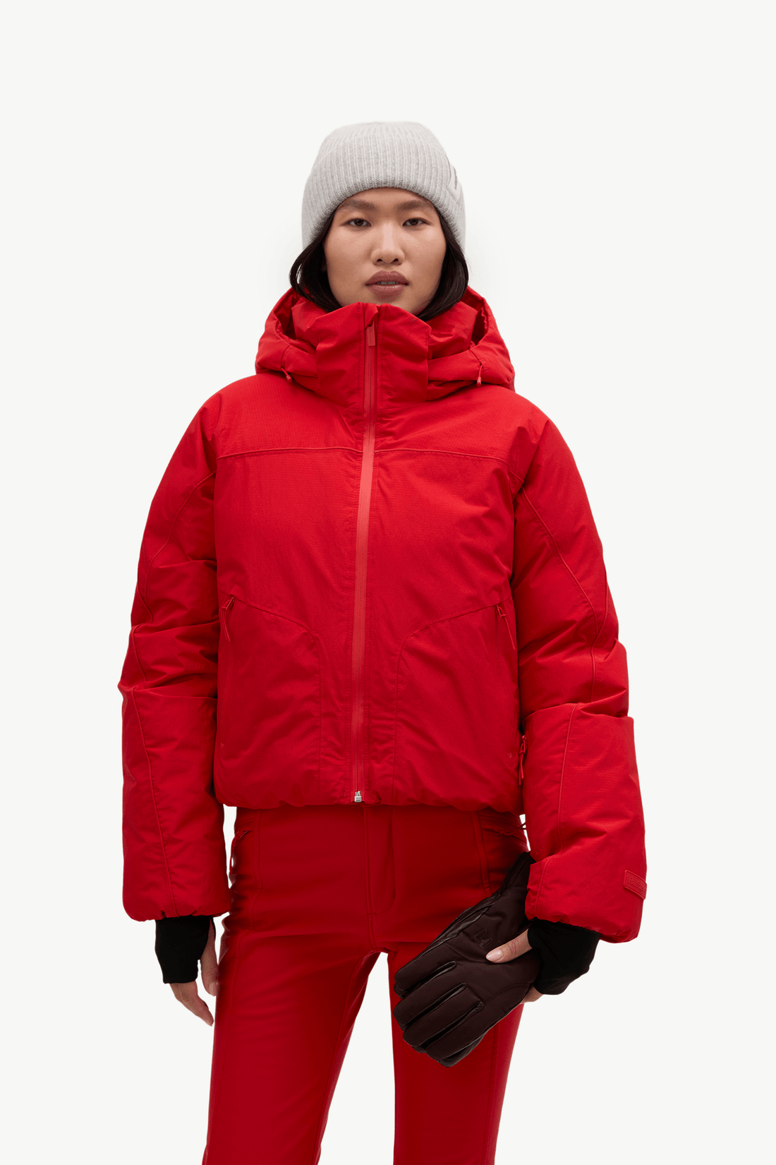 A model wearing a bright red hooded puffer jacket, matching red pants, a light gray beanie, and dark brown gloves, standing against a plain white studio background.
