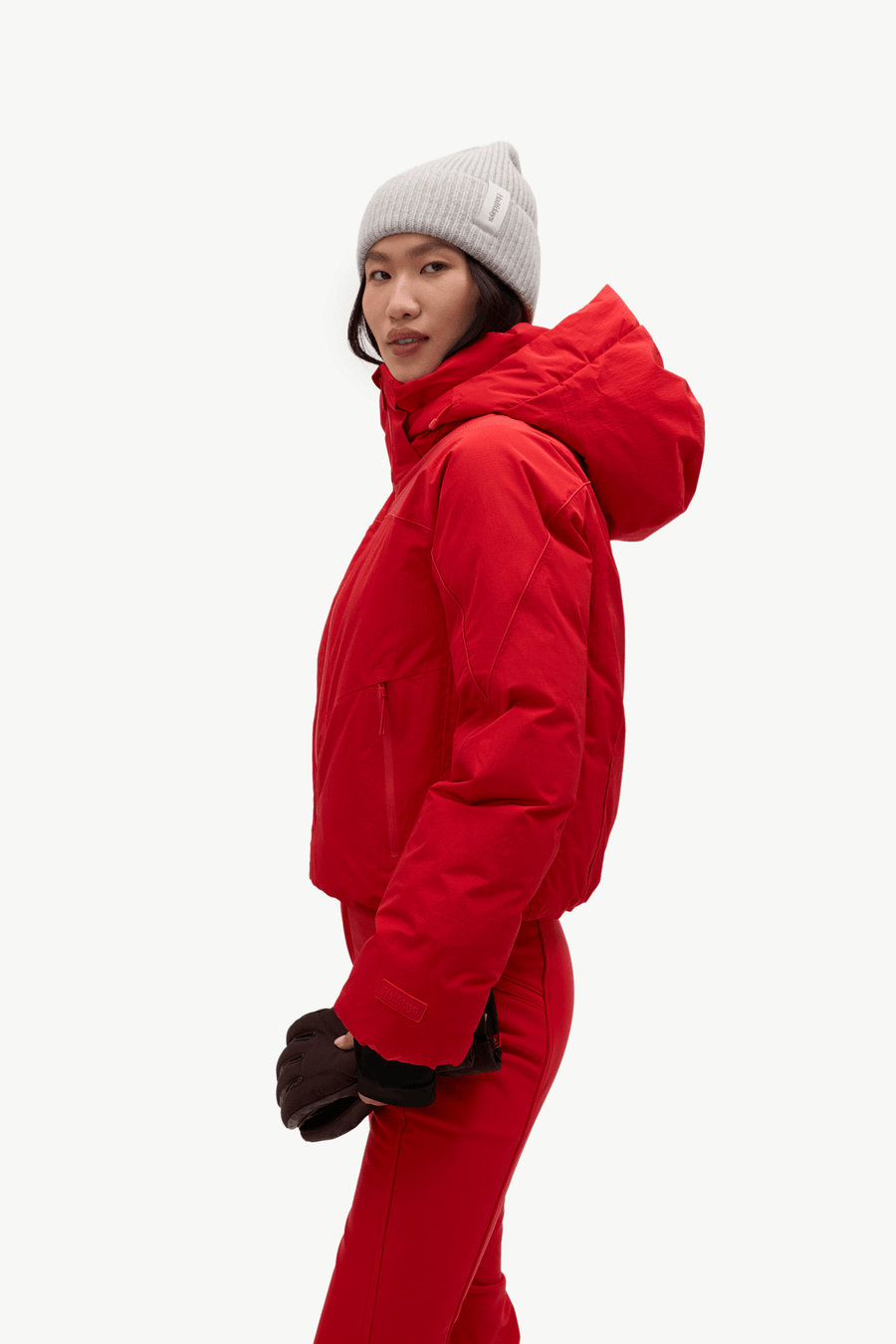 A model wearing a bright red puffer jacket, matching red pants, black gloves, and a light gray knit beanie, shown in profile against a plain white studio background.