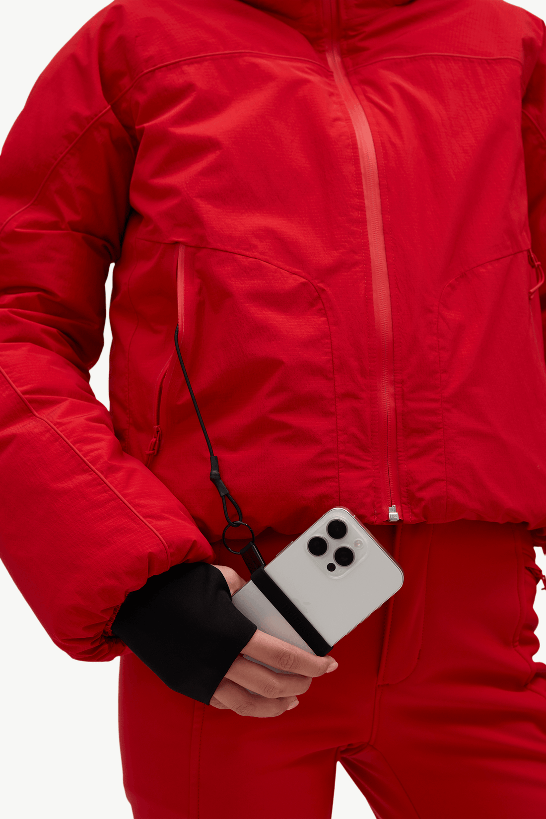 A close-up of a person wearing a bright red ski jacket and matching red pants, holding a silver smartphone with a black wrist strap attached near the pocket.