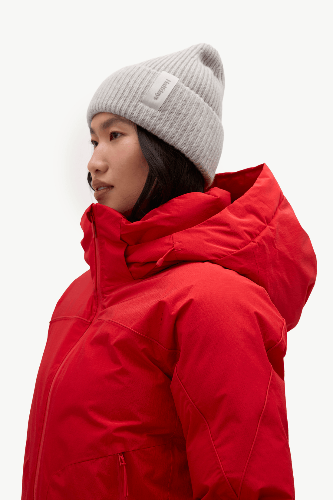 A model in profile wearing a bright red hooded winter jacket and a light gray ribbed knit beanie with a small white brand patch.