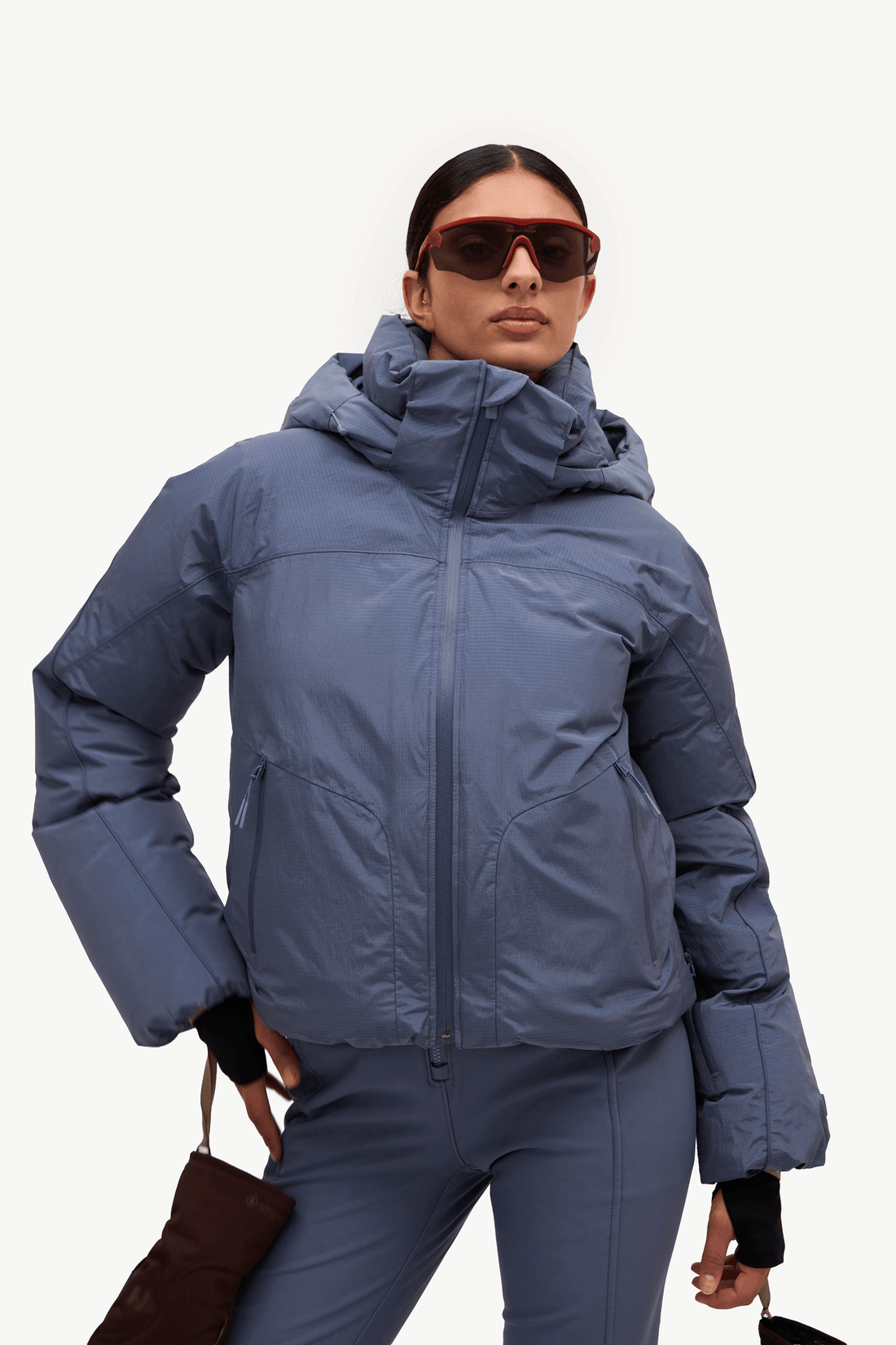 A model wearing a slate blue hooded puffer jacket and matching blue ski pants, accessorized with red-framed sunglasses and a brown leather bag, against a plain white studio background.