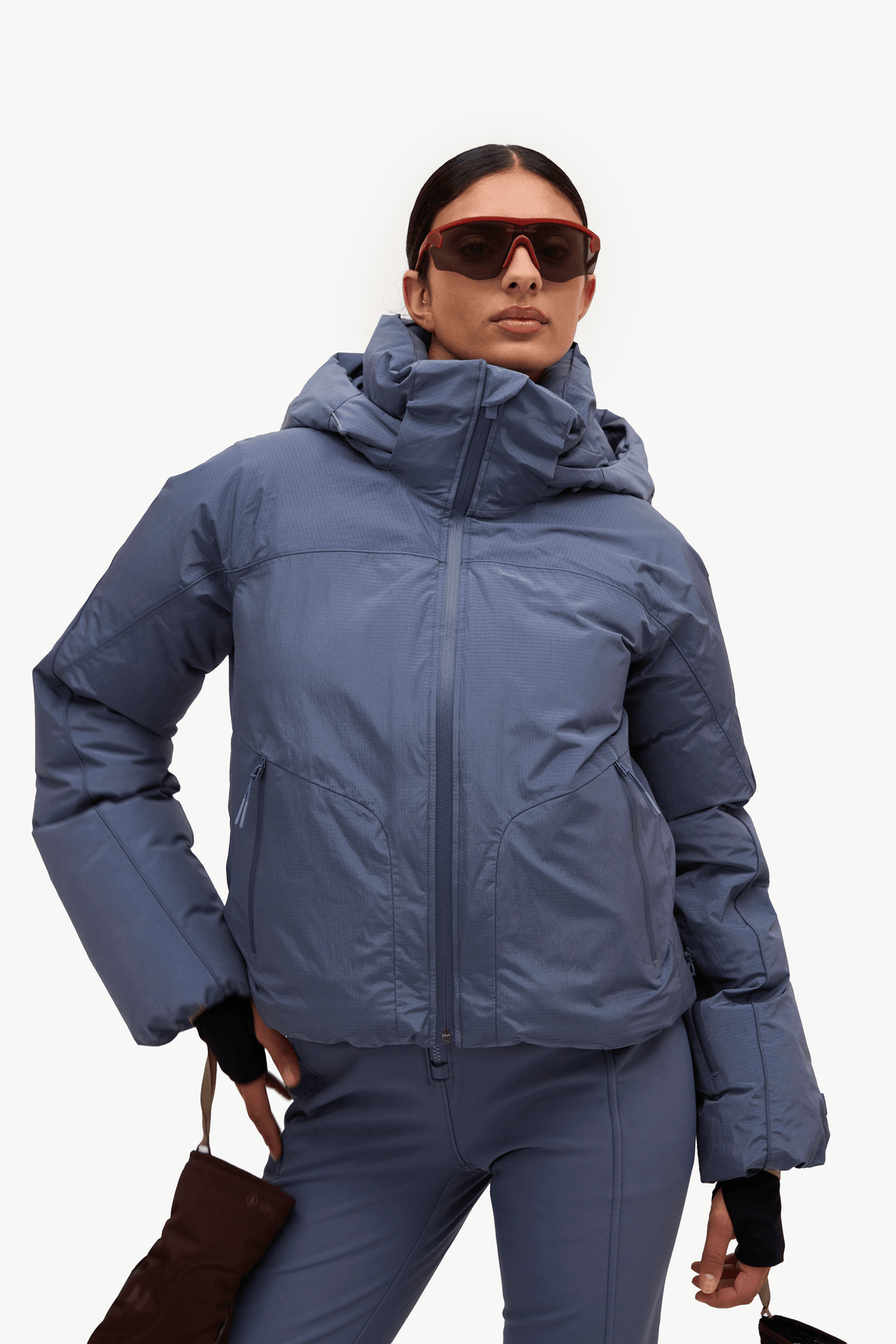 A model wearing a slate blue hooded puffer jacket and matching blue ski pants, accessorized with red-framed sunglasses and a brown leather bag, against a plain white studio background.