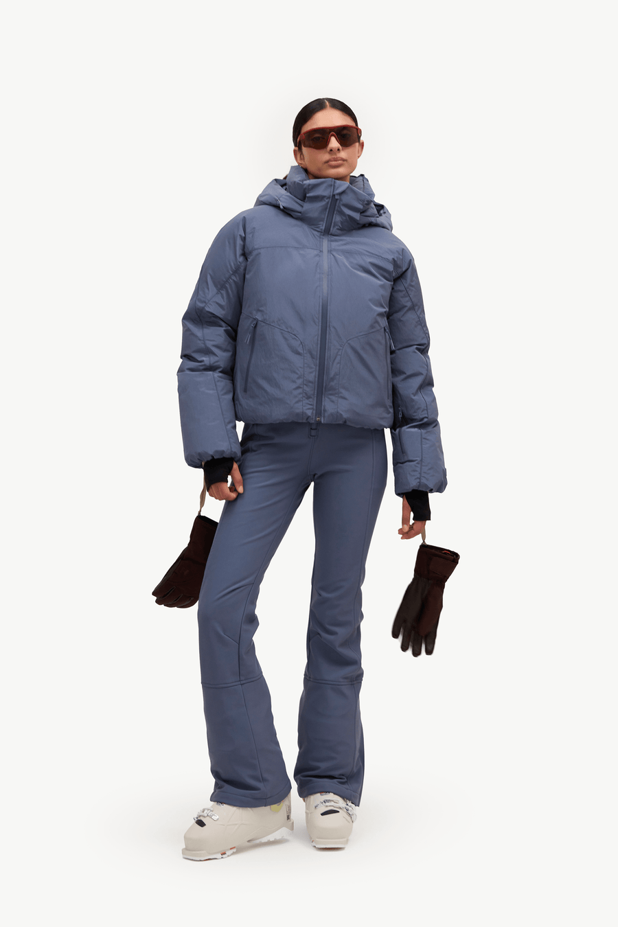A model wearing a slate blue puffer jacket, matching ski pants, and dark brown gloves, standing against a solid white studio background.