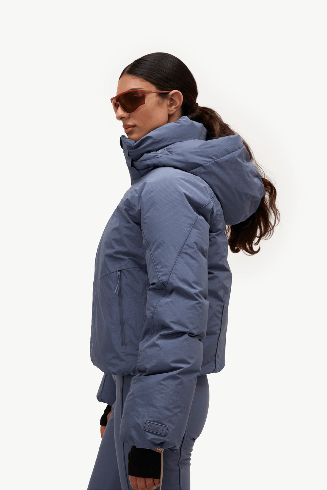 A woman in profile wearing a slate blue puffer jacket and matching pants against a plain white studio background.