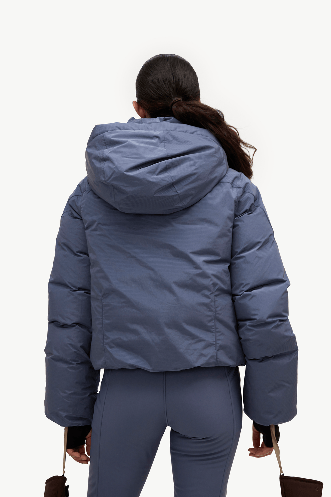 A woman viewed from behind wearing a slate blue puffer jacket and matching blue leggings against a plain white studio background.