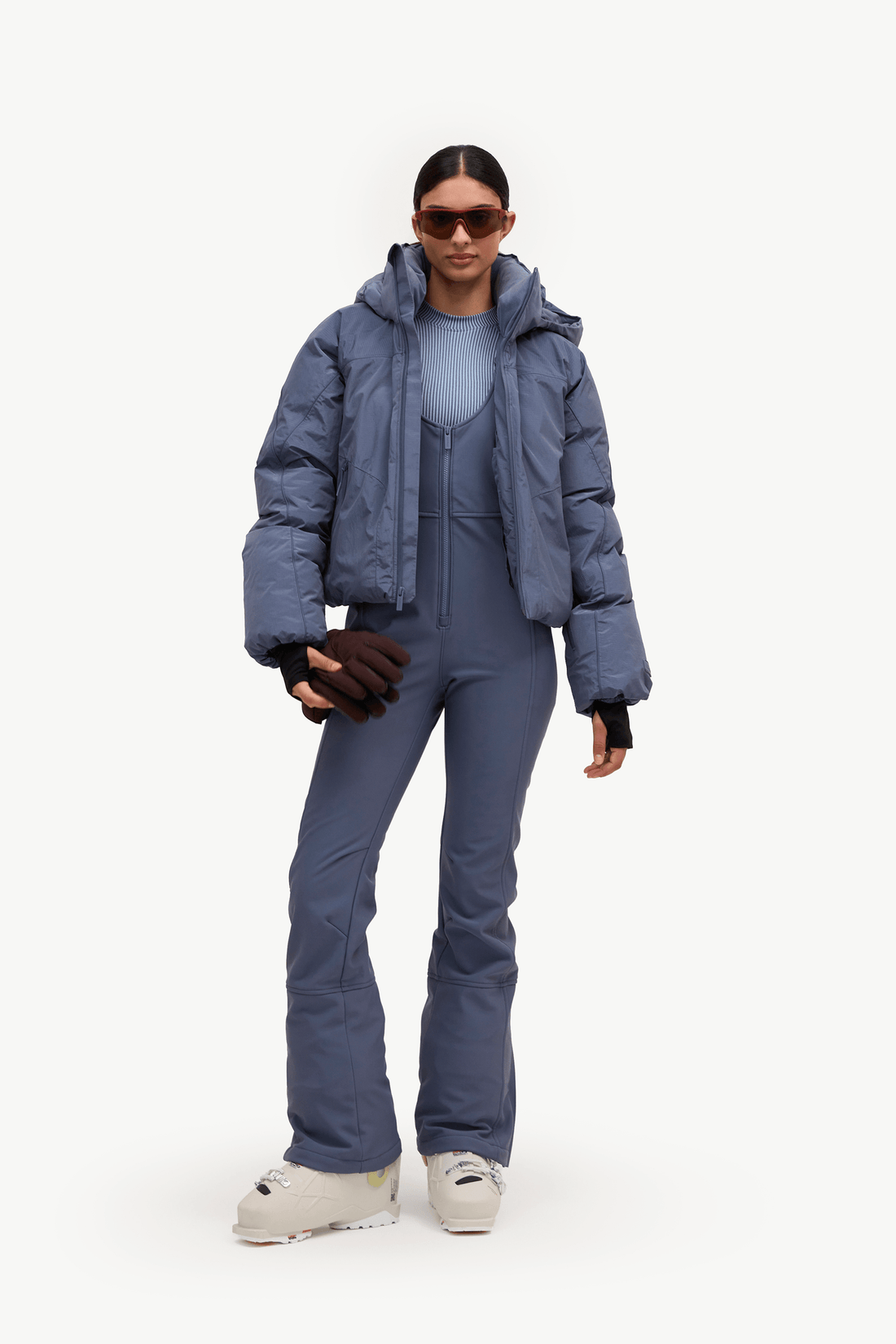 A model wearing a slate blue ski outfit consisting of a puffer jacket, a matching zip-up hoodie, and ski pants, paired with white ski boots and sunglasses, against a plain white studio background.