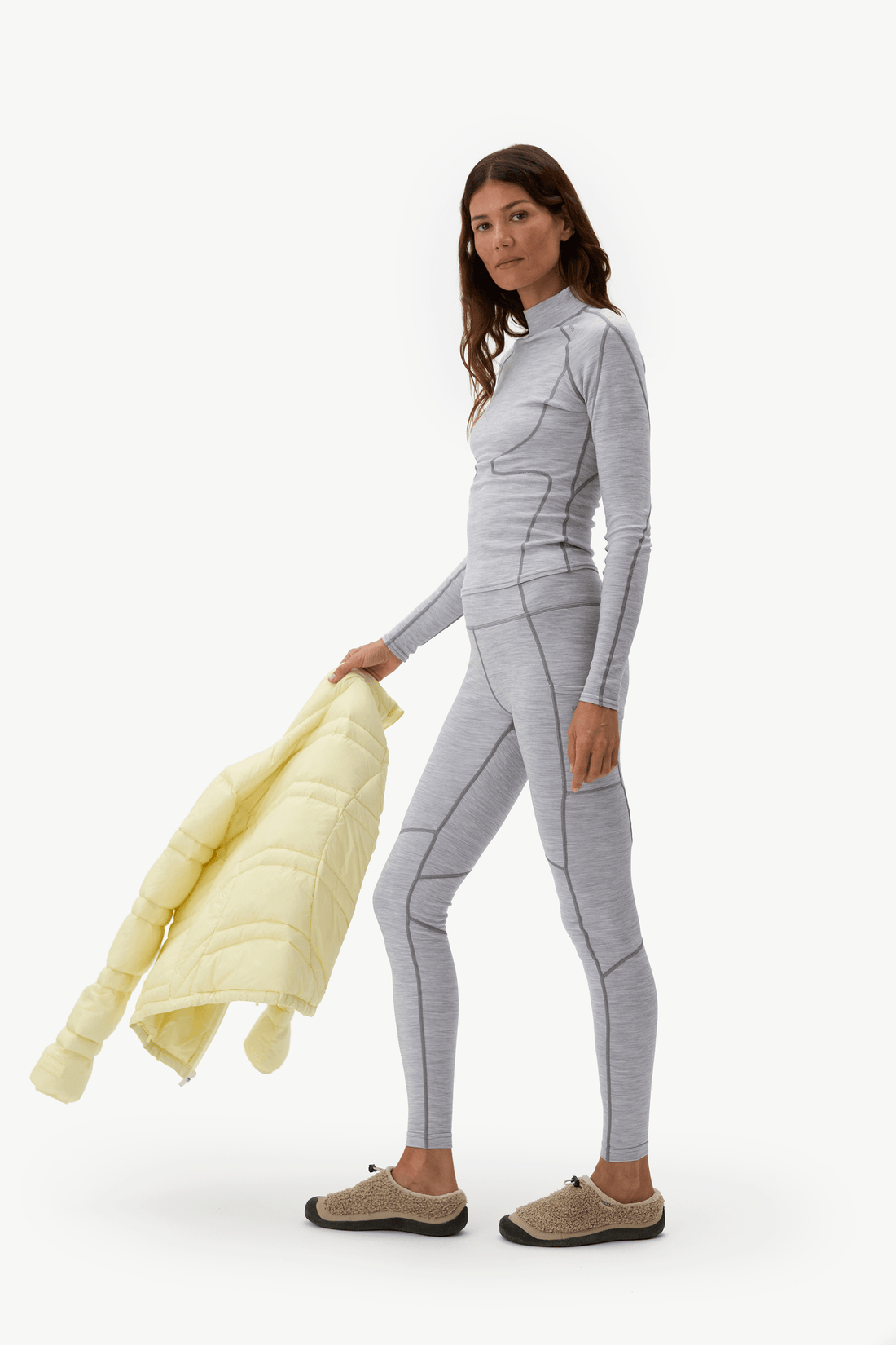 A woman modeling a light gray long-sleeve athletic one-piece suit, holding a pale yellow puffer jacket against a plain white studio background.