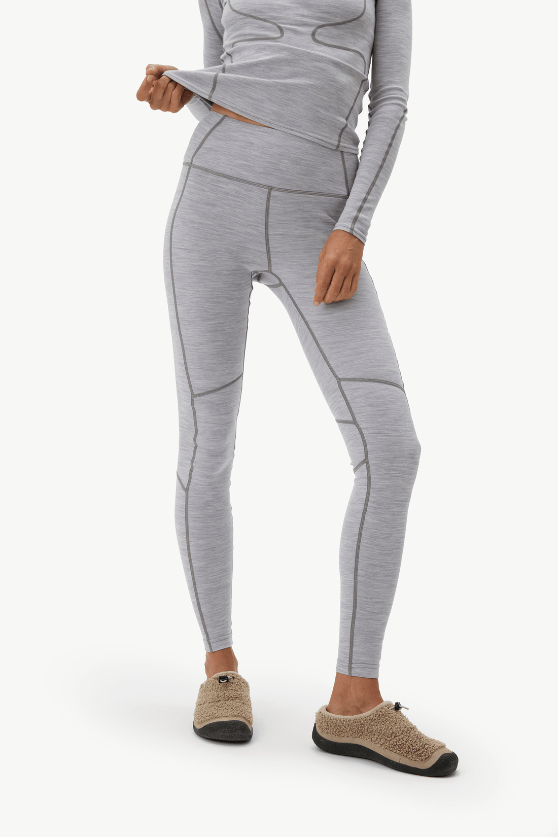 A model wearing light gray heathered athletic leggings with contrast darker gray seam lines, paired with a matching long-sleeve top and tan slip-on shoes, shown against a plain white studio background.