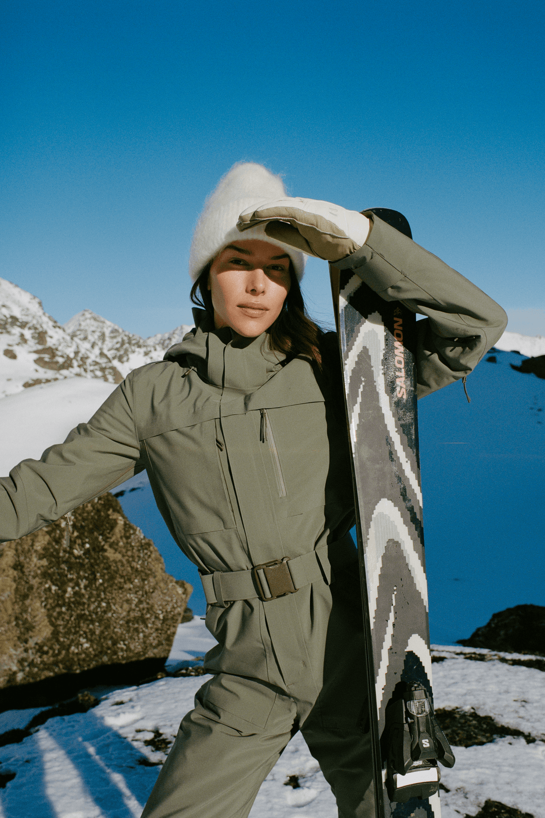 A woman in a snowy mountain setting holds a snowboard while wearing an olive green ski suit, white beanie, and white gloves. The background features a clear blue sky and snow-covered peaks.