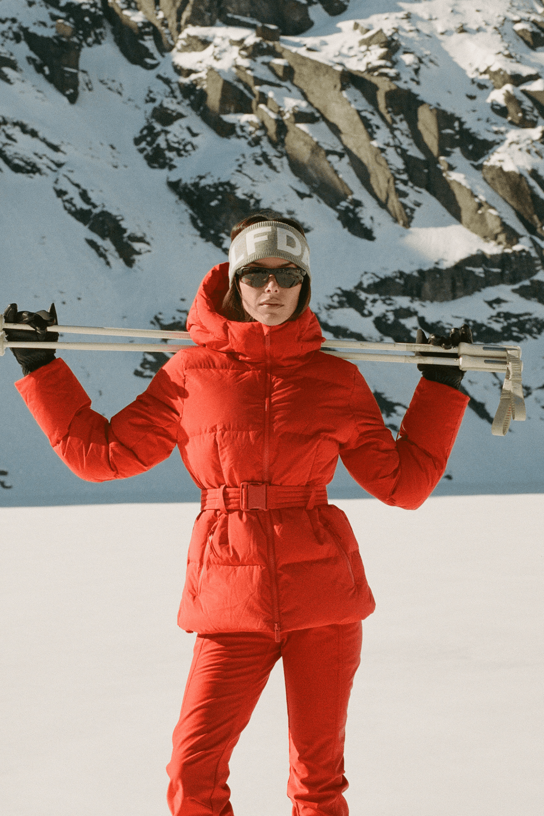 A person standing in a snowy mountain setting wearing a bright red puffer jacket and matching red ski pants, with ski poles across their shoulders and a white headband.