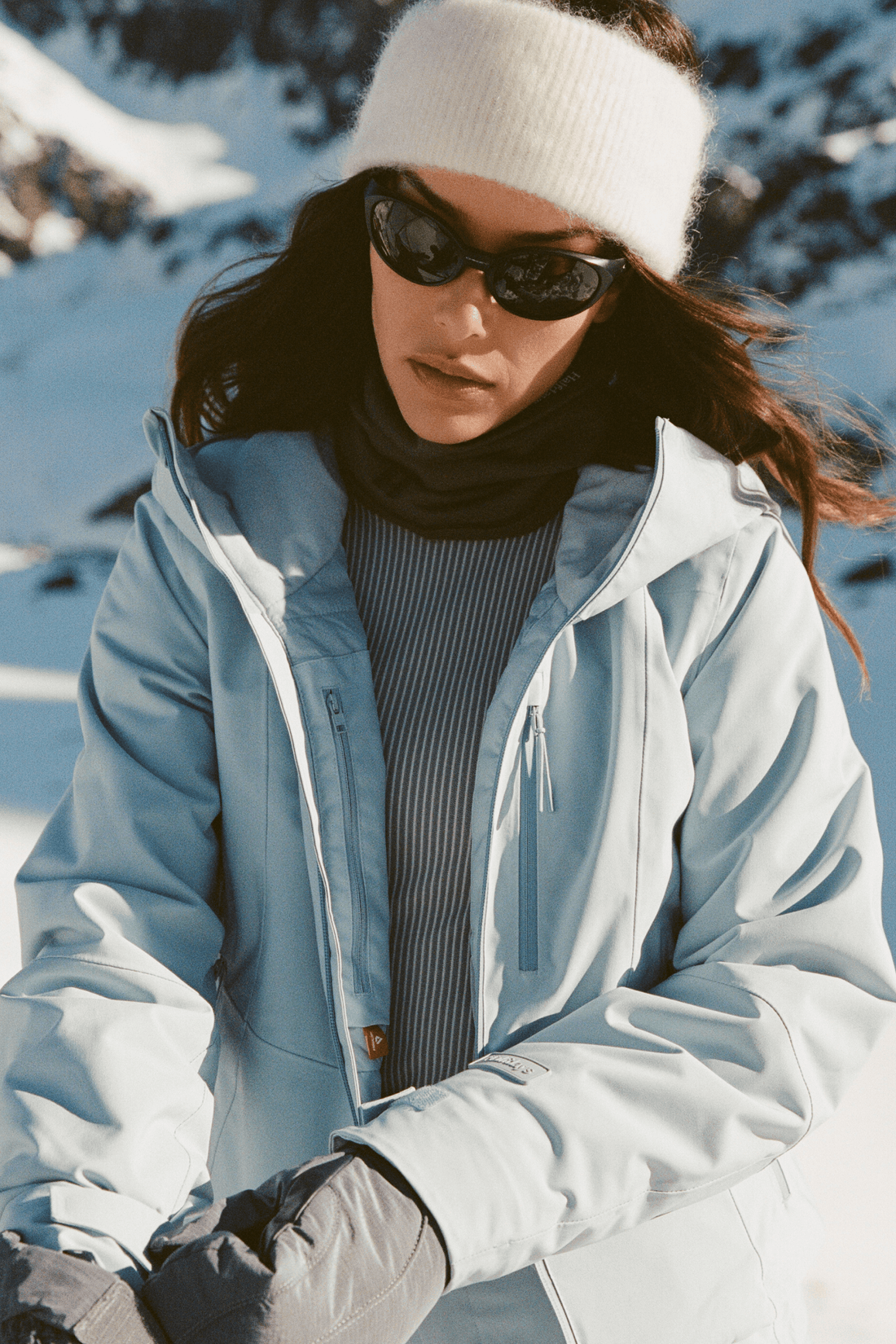 A woman in a snowy outdoor setting wearing a light blue ski jacket, a white knit beanie, black sunglasses, a dark ribbed turtleneck, and gray gloves.