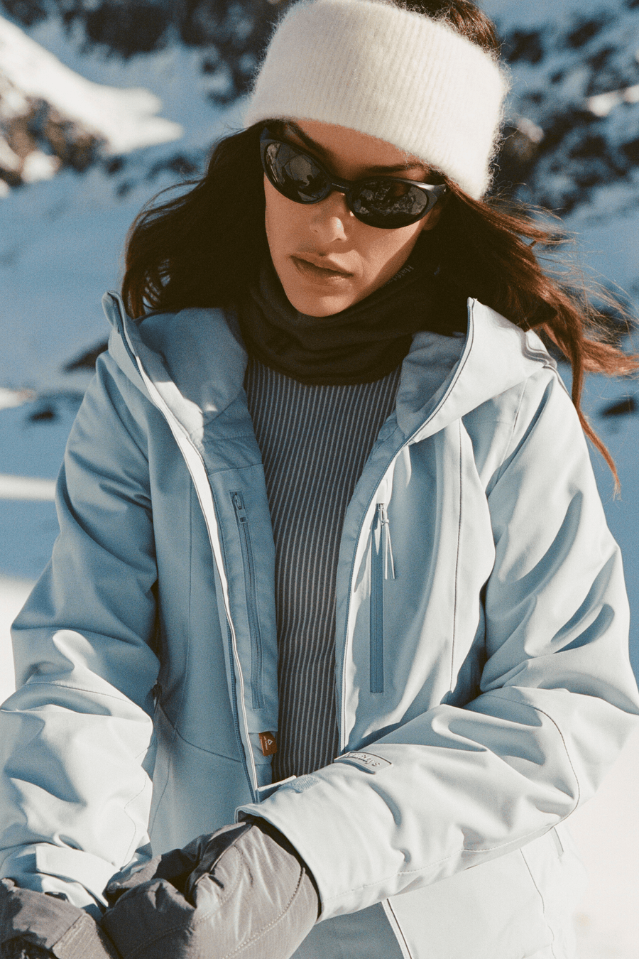 A woman in a snowy outdoor setting wearing a light blue ski jacket, a white knit beanie, black sunglasses, a dark ribbed turtleneck, and gray gloves.