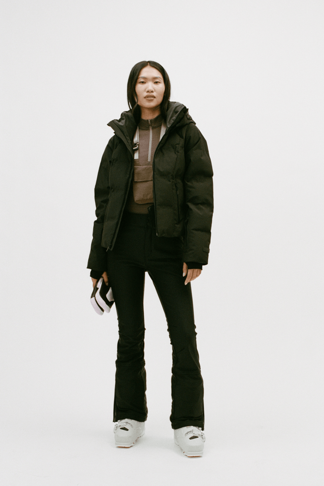 A model stands against a plain light gray studio background wearing a dark olive green puffer jacket over a brown top, black pants, and white sneakers, holding a small black-and-white item in one hand.
