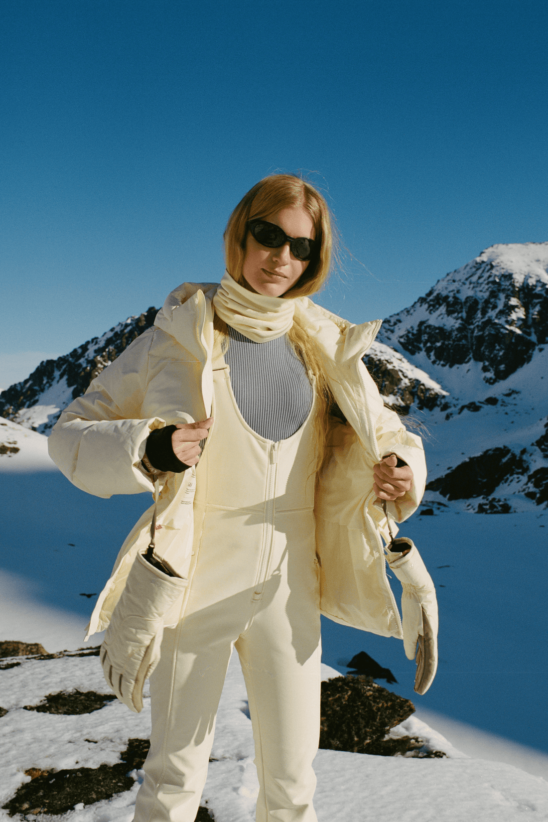 A woman wearing a cream-colored ski suit and matching gloves stands in a snowy mountain landscape under a clear blue sky, holding her ski jacket open.