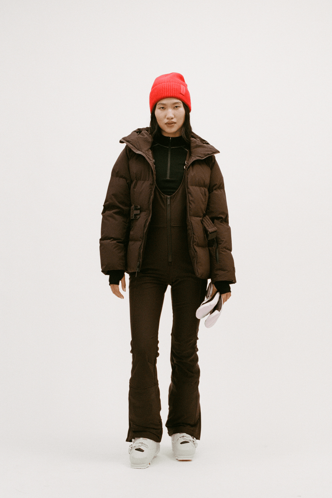 A model stands against a light gray studio background wearing a dark brown puffer jacket, matching dark brown pants, a black top, and a bright red knit beanie, holding a pair of white gloves.