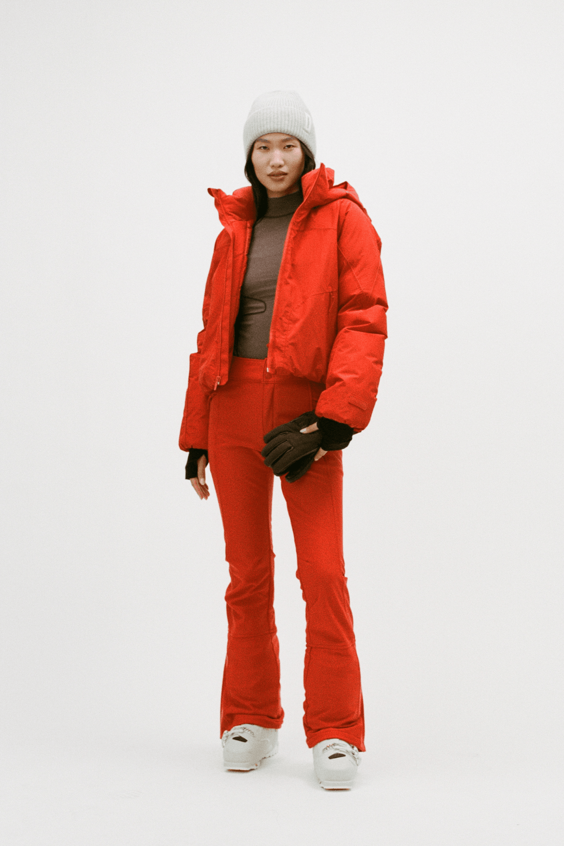 A model stands against a light gray studio background wearing a bright red puffer jacket and matching red pants, with a white beanie, black gloves, a brown top, and white sneakers.