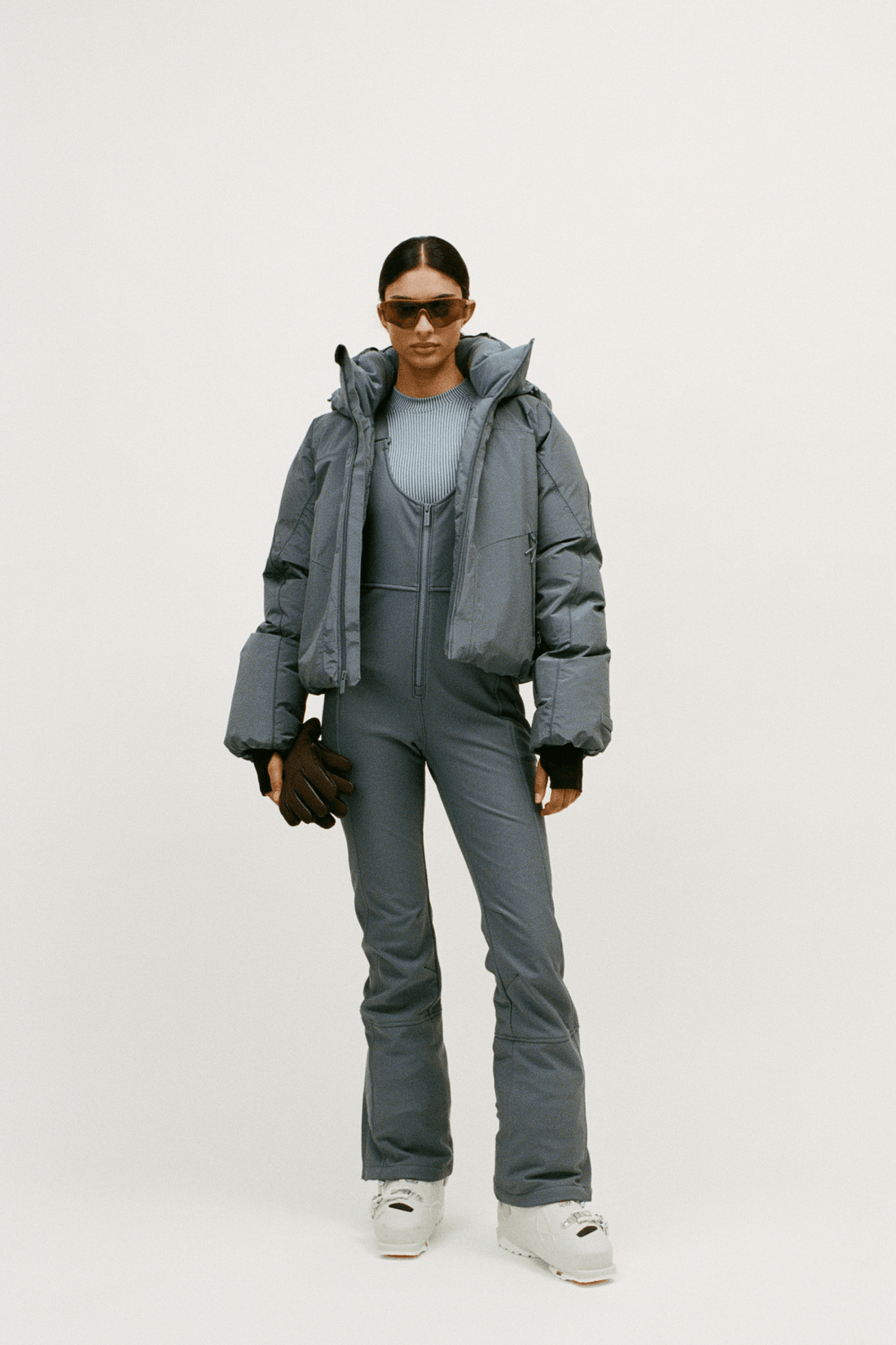 A model stands against a light gray studio background wearing a gray puffer jacket over a matching gray one-piece jumpsuit, with white sneakers and dark sunglasses.