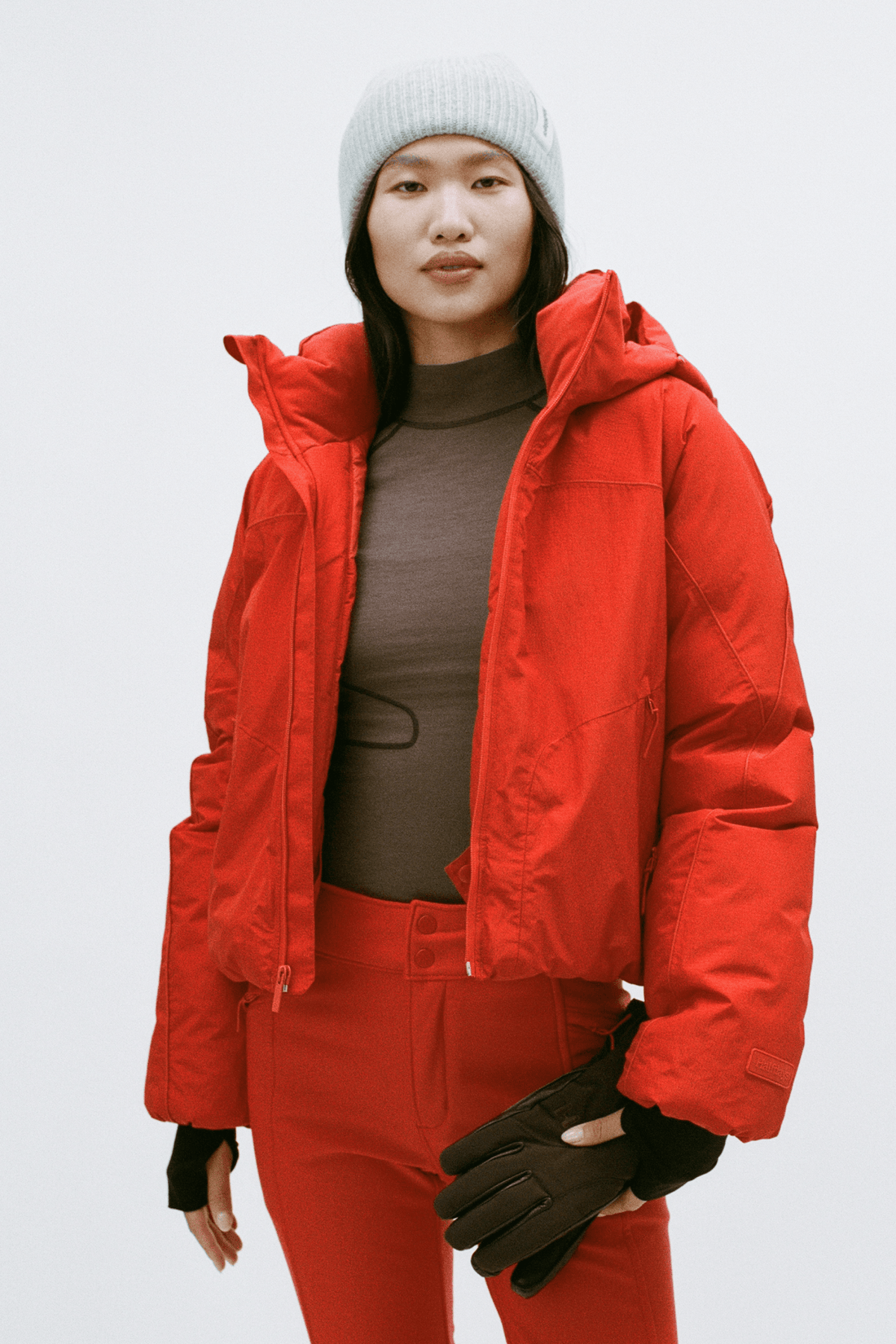 A model wearing a bright red hooded puffer jacket, matching red pants, a brown ribbed top, black gloves, and a light gray beanie against a plain light gray studio background.