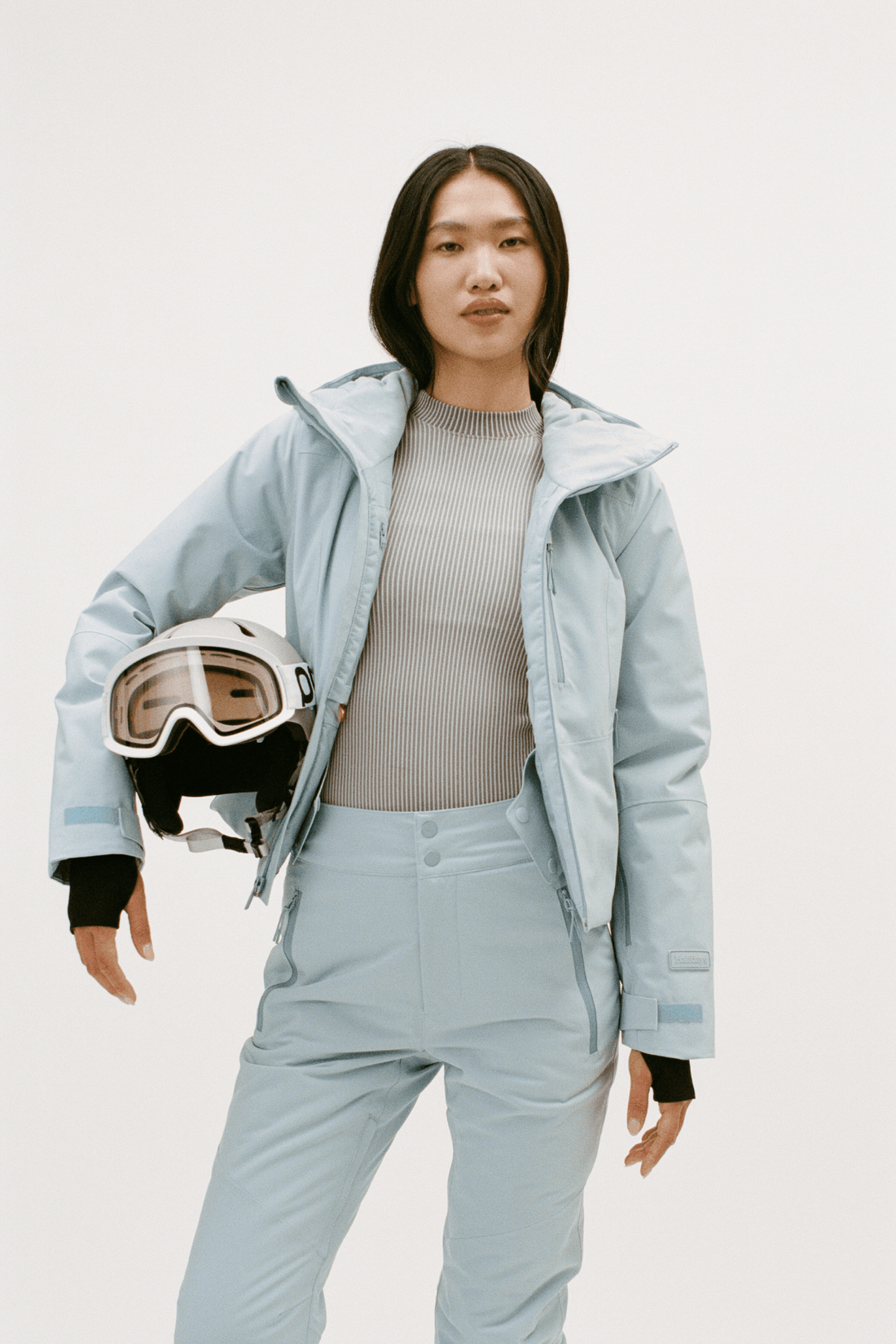 A model wearing a light blue ski jacket and matching ski pants, holding a white ski helmet with goggles, against a plain white studio background.