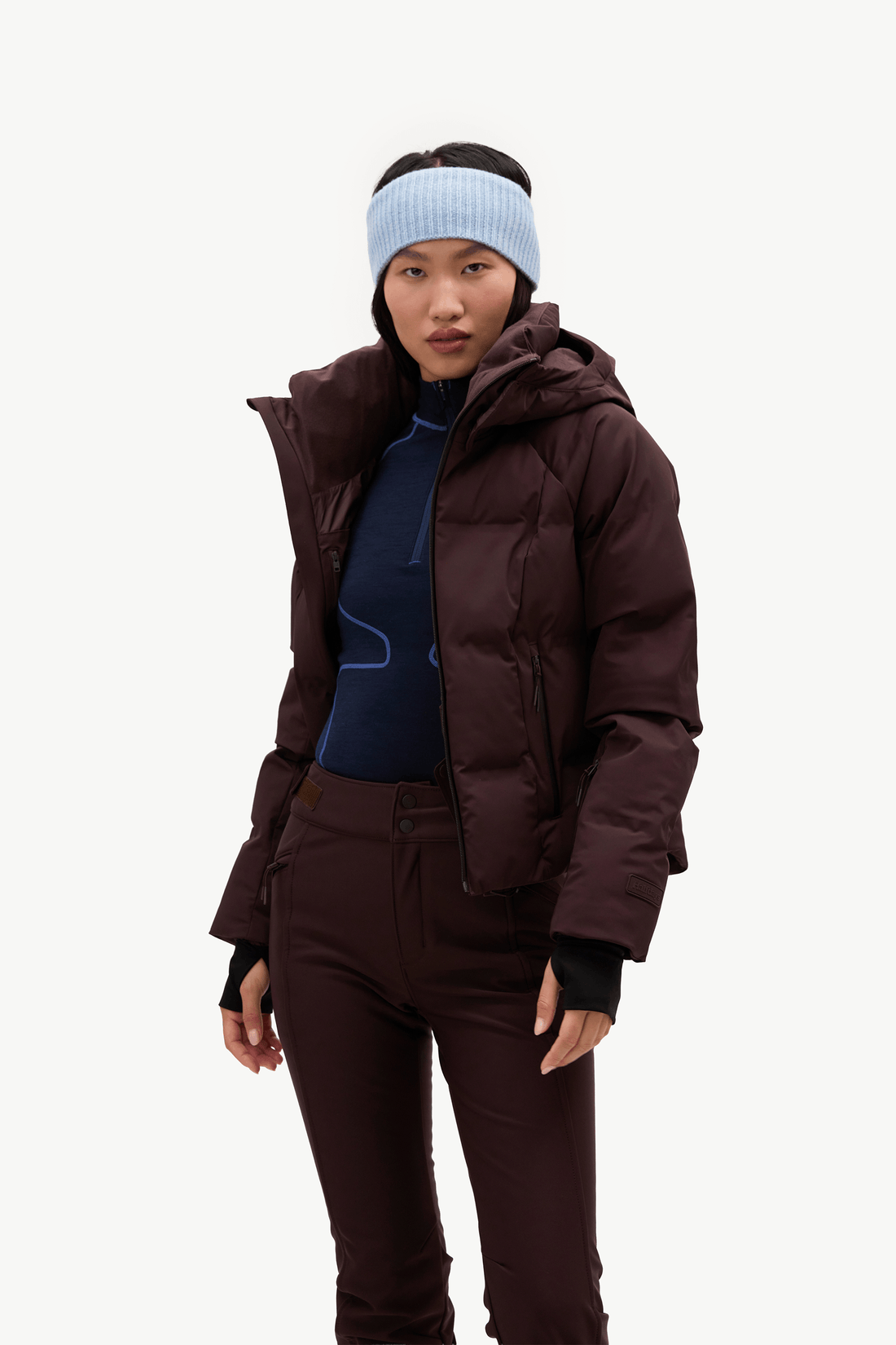 A model wearing a dark burgundy hooded puffer jacket, matching dark pants, a light blue knit beanie, against a clean white background.