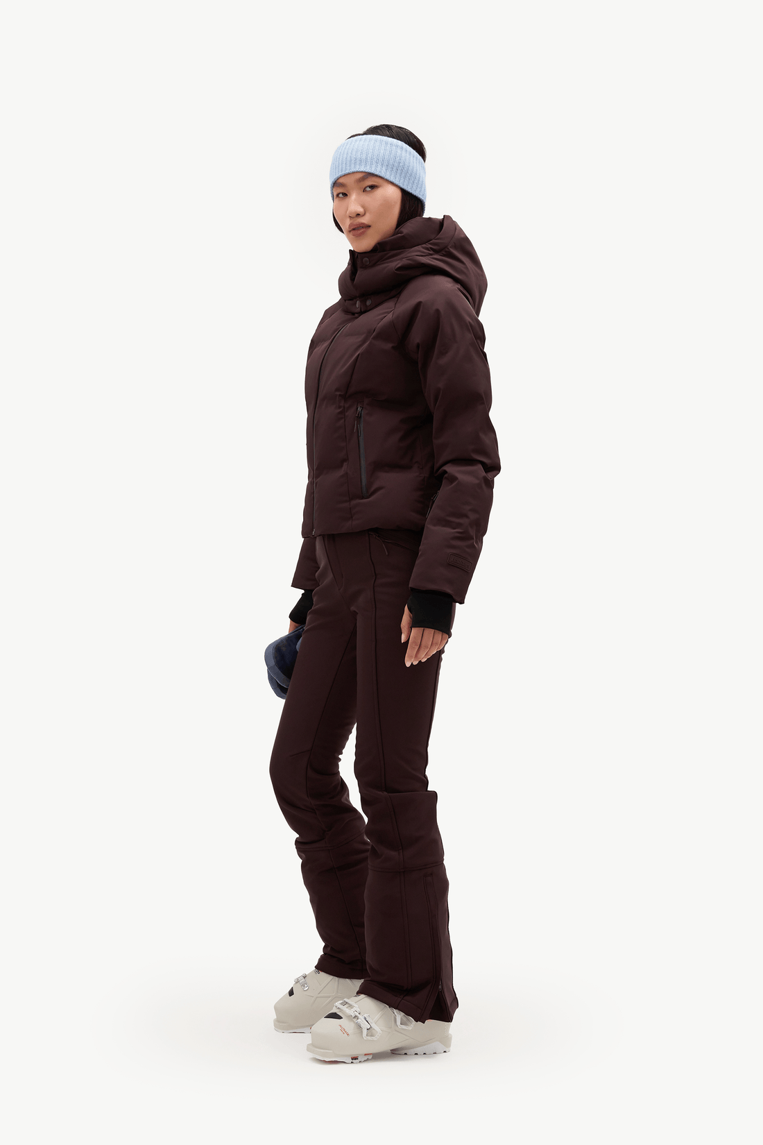 A model wearing a dark burgundy hooded puffer jacket, matching dark pants, a light blue knit beanie, against a clean white background.