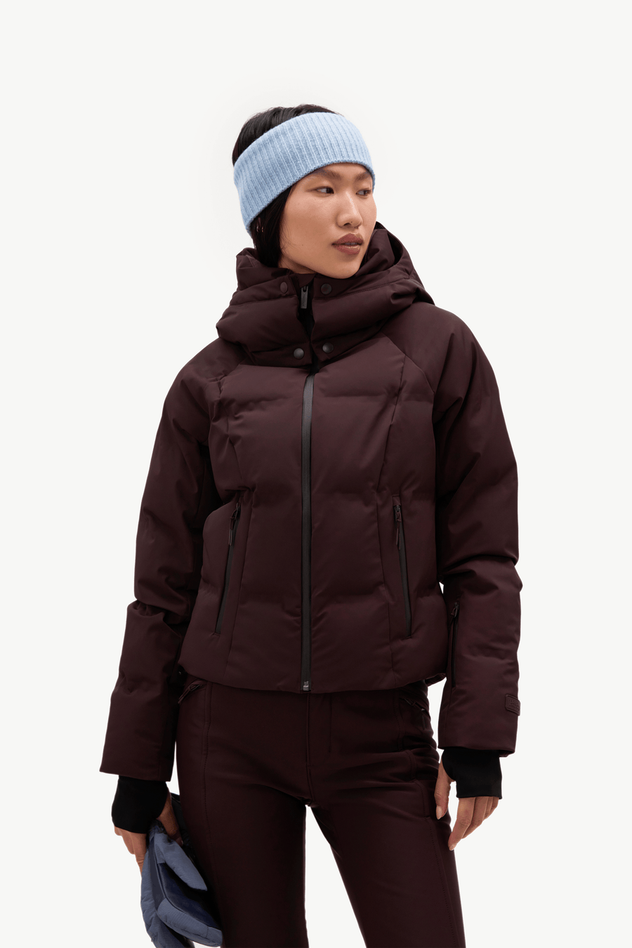 A model wearing a dark burgundy hooded puffer jacket, matching dark pants, a light blue knit beanie, and holding a blue item against a clean white background.