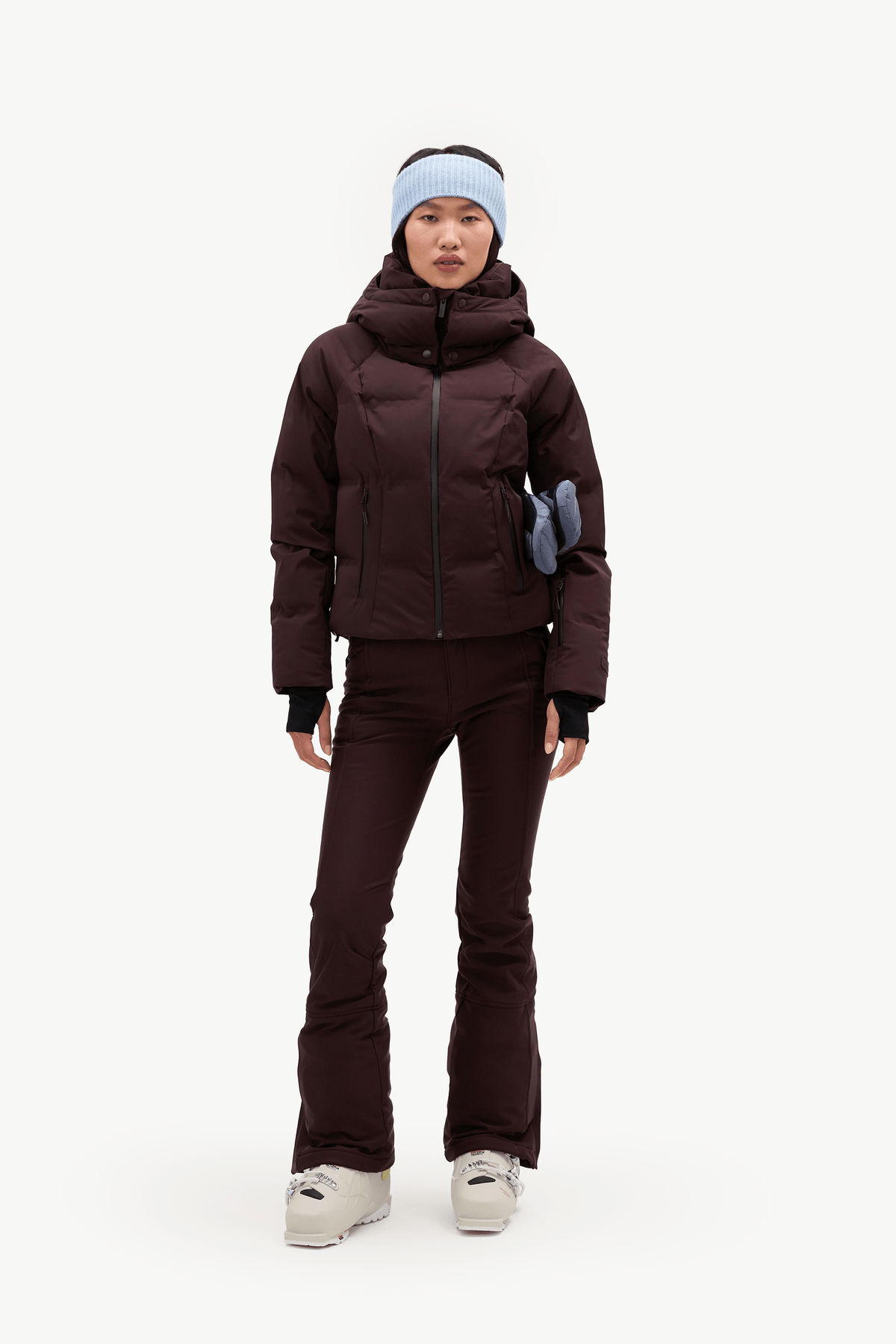 A model wearing a dark burgundy hooded puffer jacket, matching dark pants, a light blue knit beanie, and holding a blue item against a clean white background.